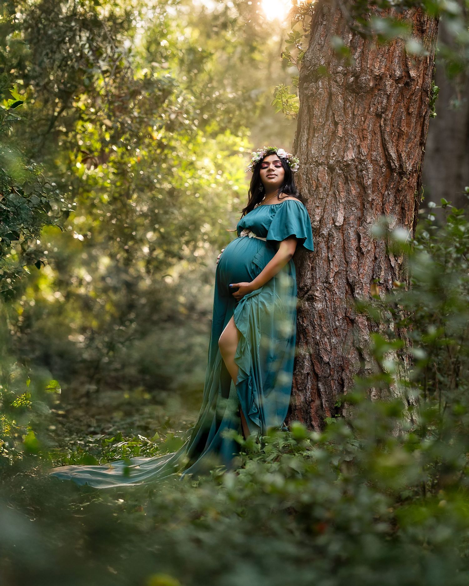 A figure in a teal dress poses gracefully against a large tree trunk in a sunlit forest setting.
