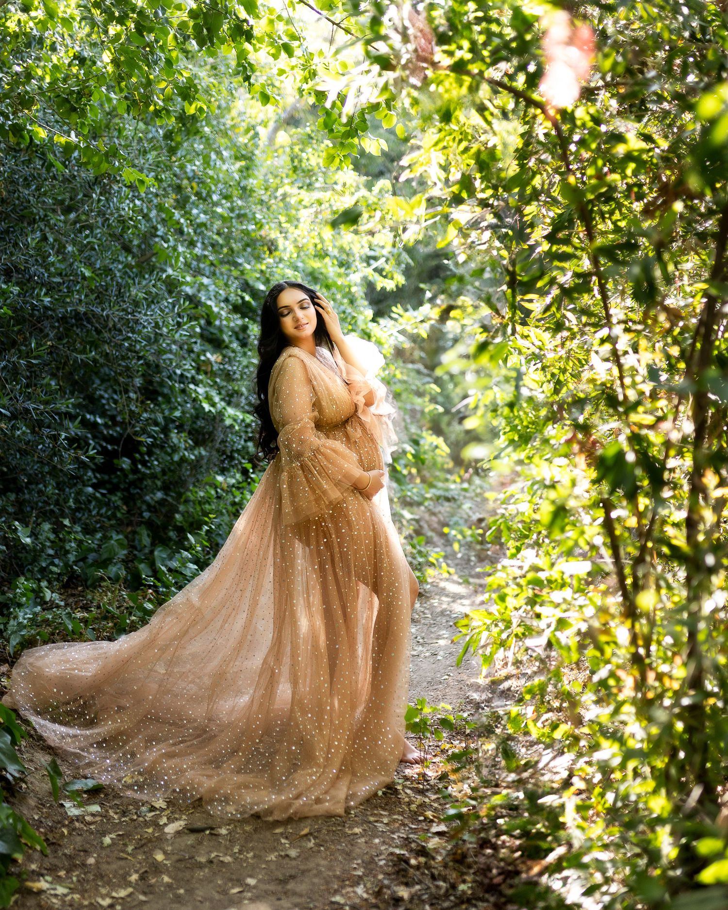 A flowing champagne-colored gown with tulle details sparkles in a sunlit forest path creating an ethereal scene.