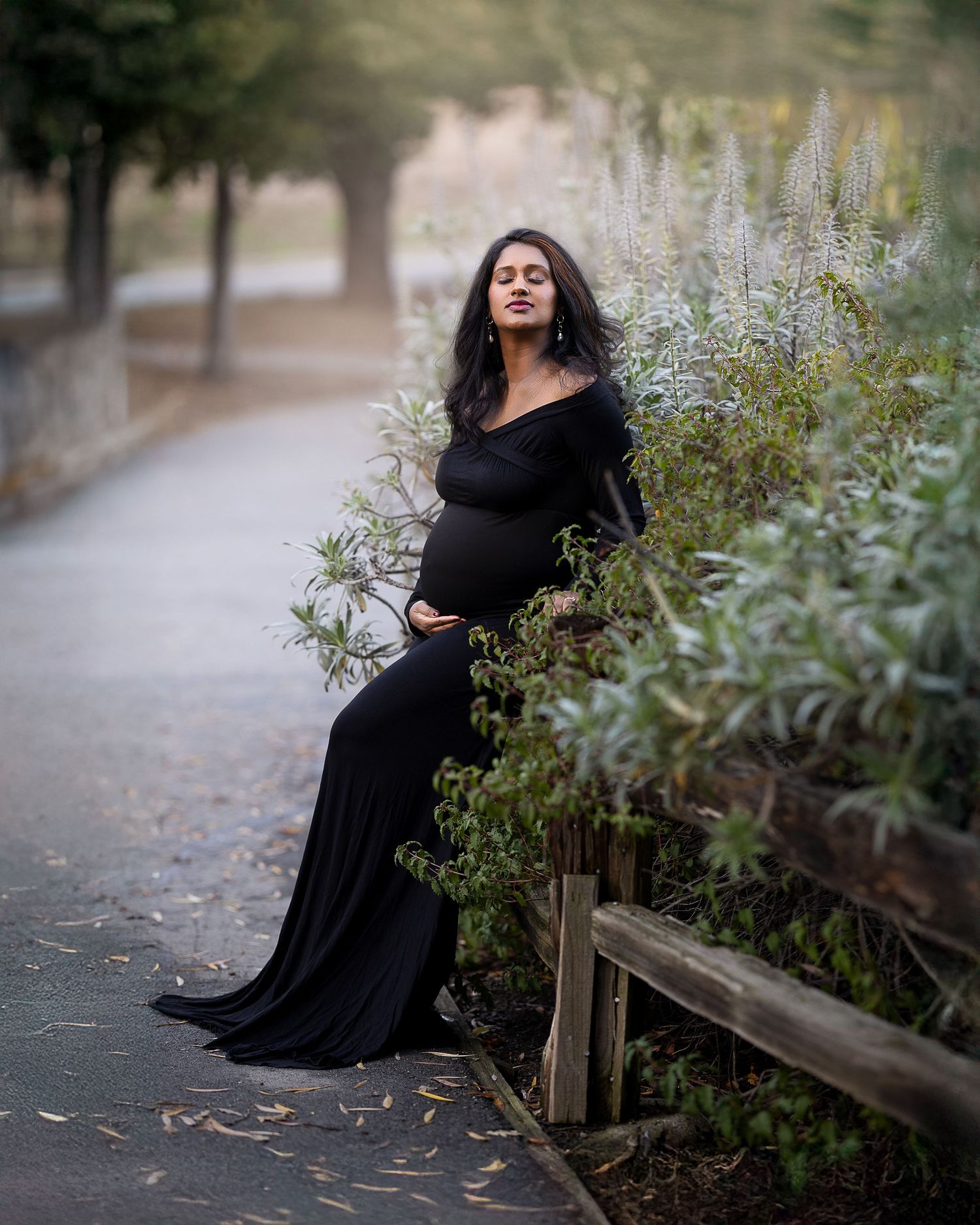 A woman in a flowing black dress poses gracefully along a wooden fence in a garden setting.