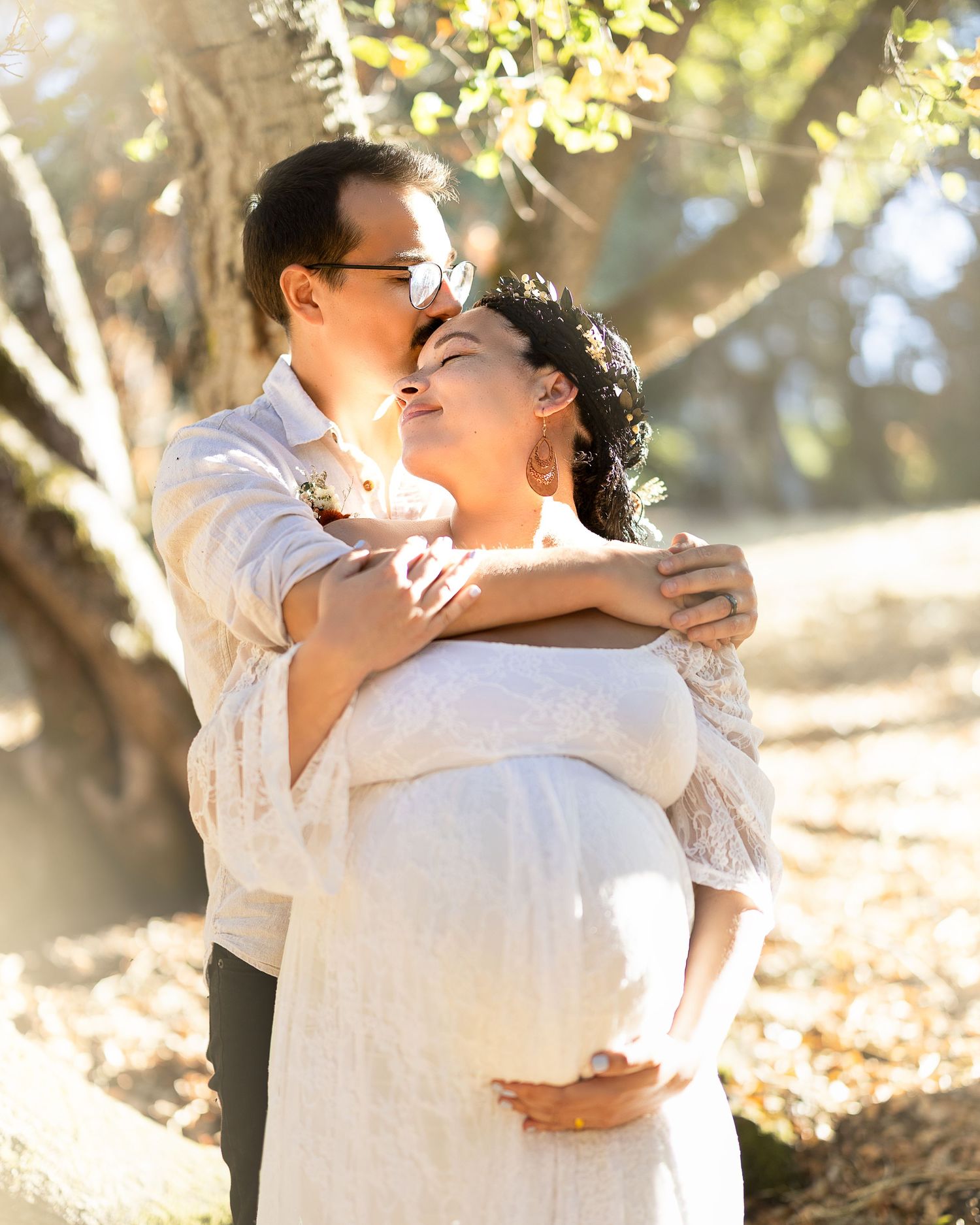 A couple in white outfits embraces lovingly in a sun-dappled outdoor setting with autumn trees in the background.