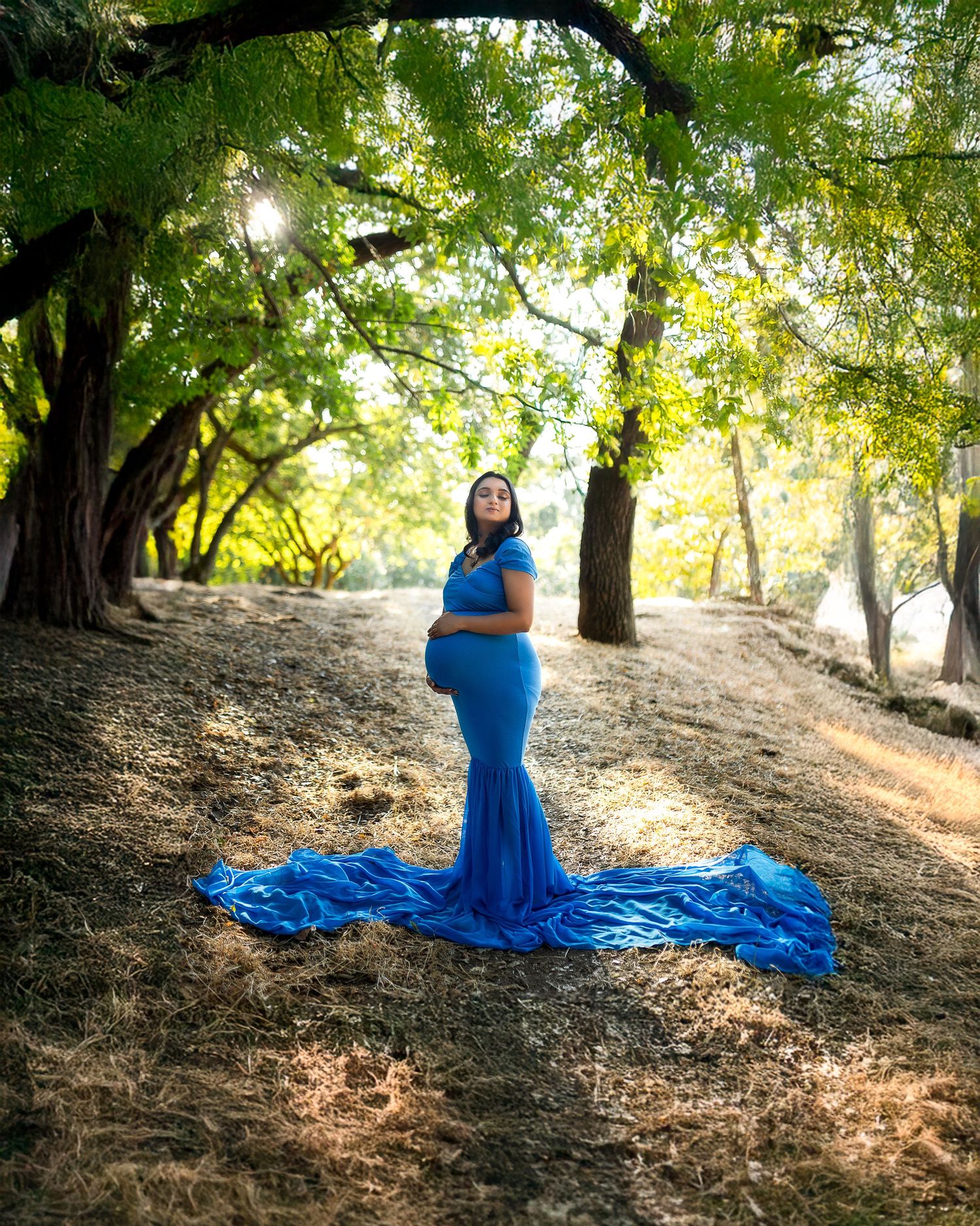 A figure in a flowing blue dress poses gracefully on a sunlit hillside among trees creating an ethereal outdoor portrait.