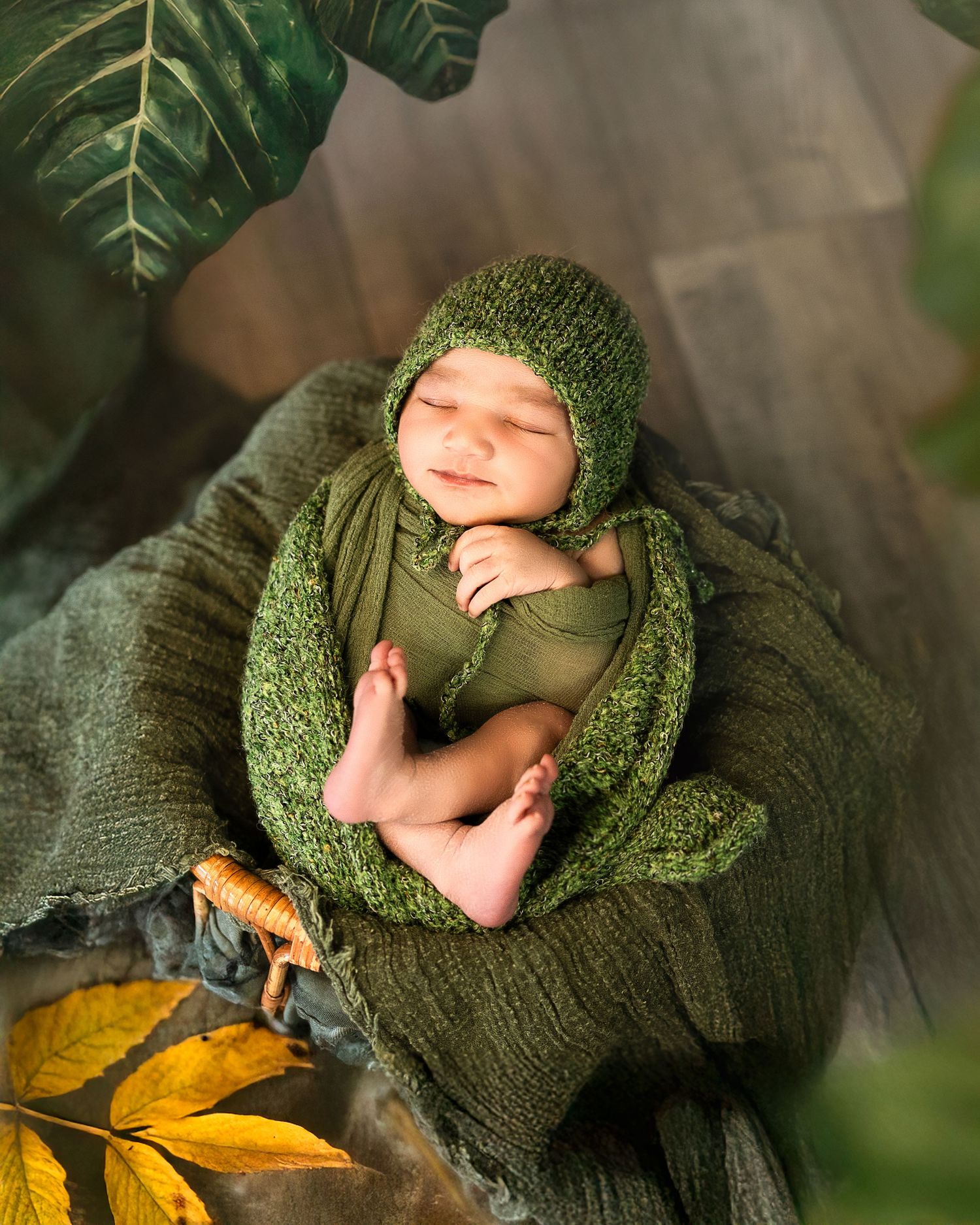 Sleeping newborn wrapped in green knit blanket with autumn leaves on gray textured background.