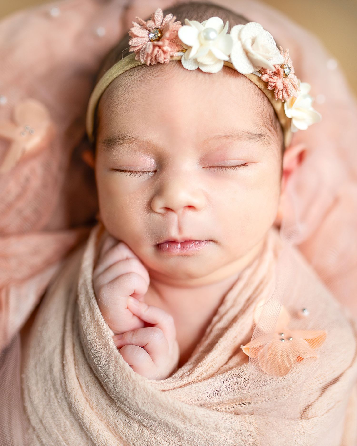 A sleeping newborn swaddled in beige fabric wearing delicate pink and white floral headbands in a peaceful pose.