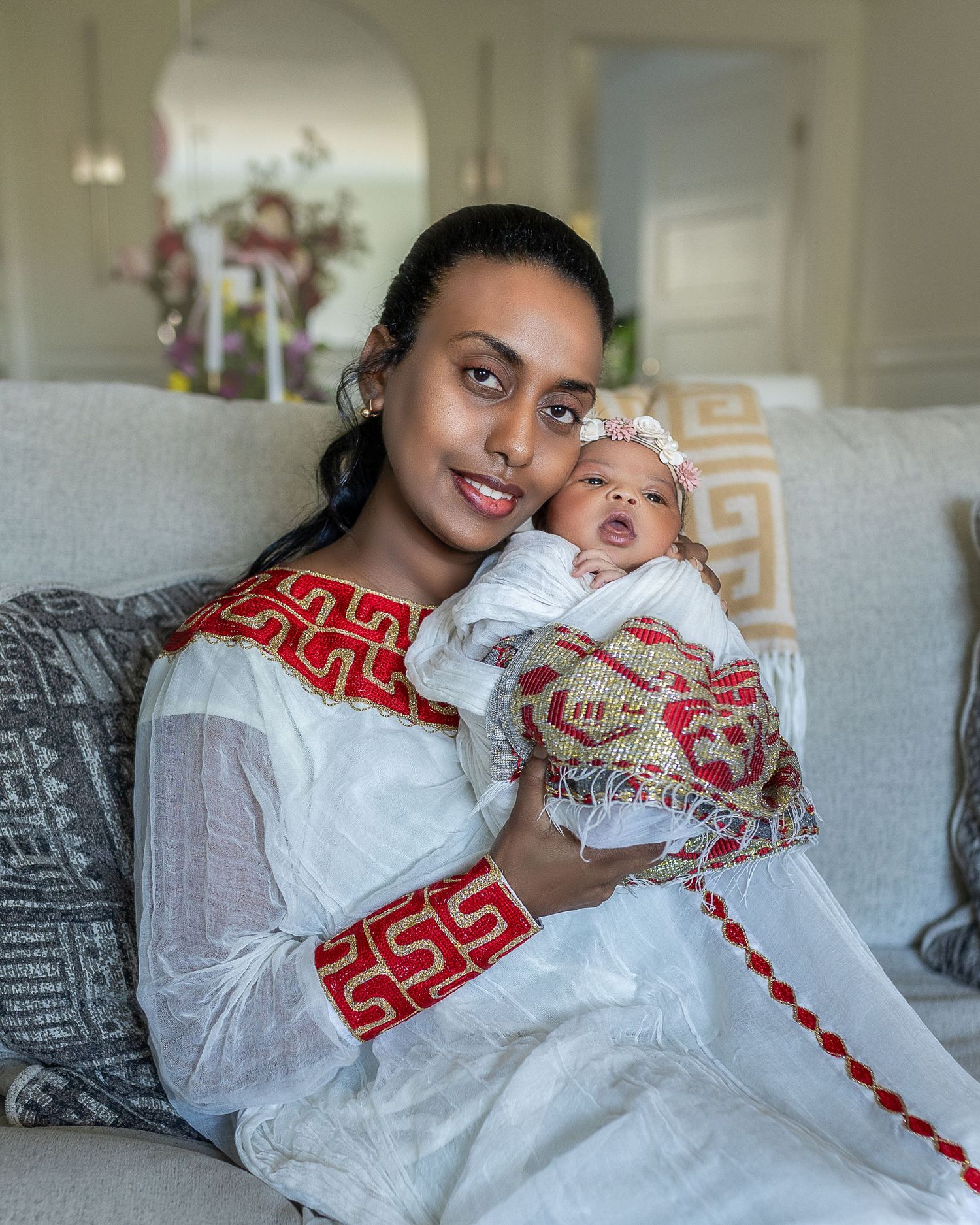 A parent and baby wearing matching white and red traditional Ethiopian dresses sit together on a gray couch.