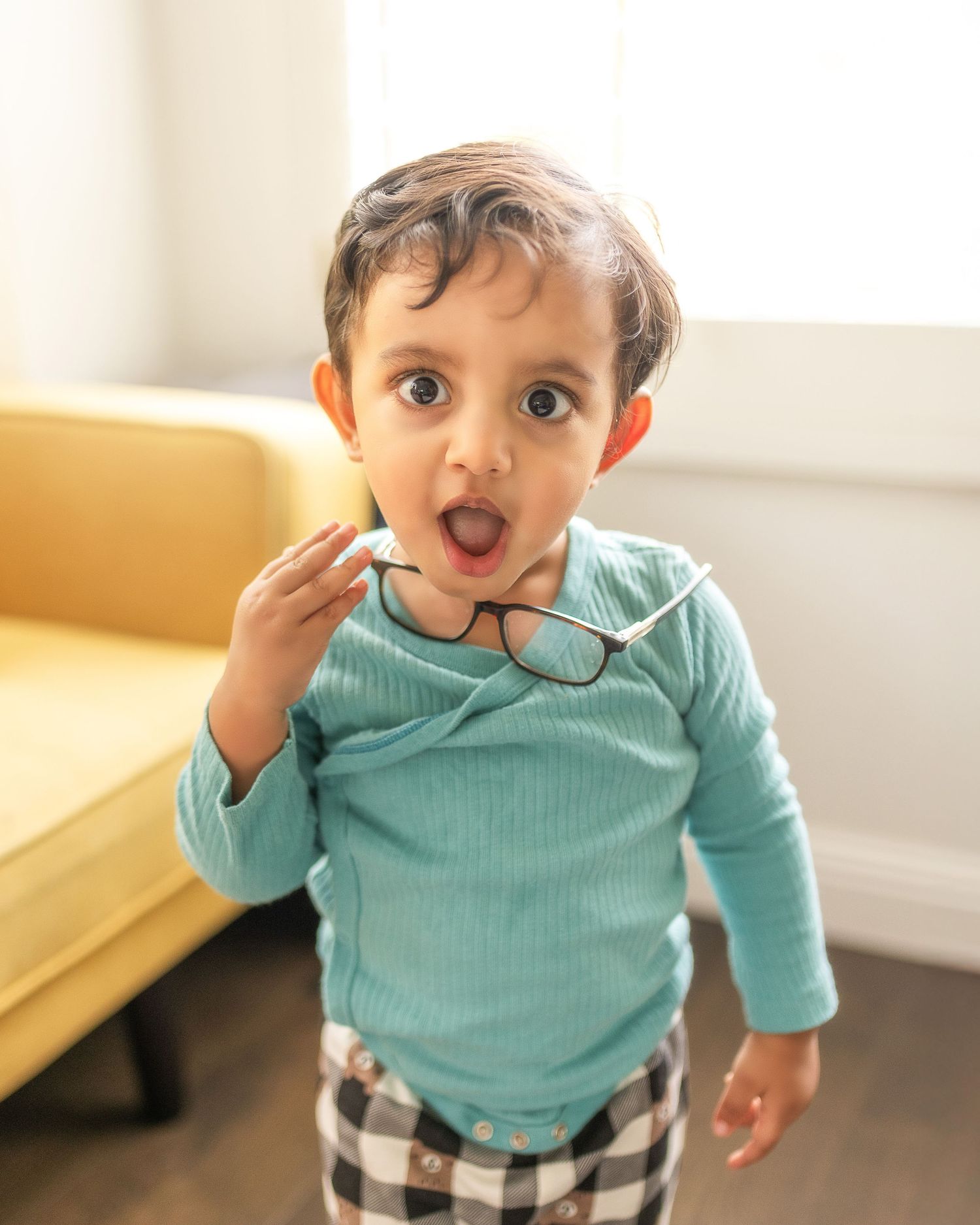 Small child in a turquoise shirt makes silly expressions while playing in a sunlit room.