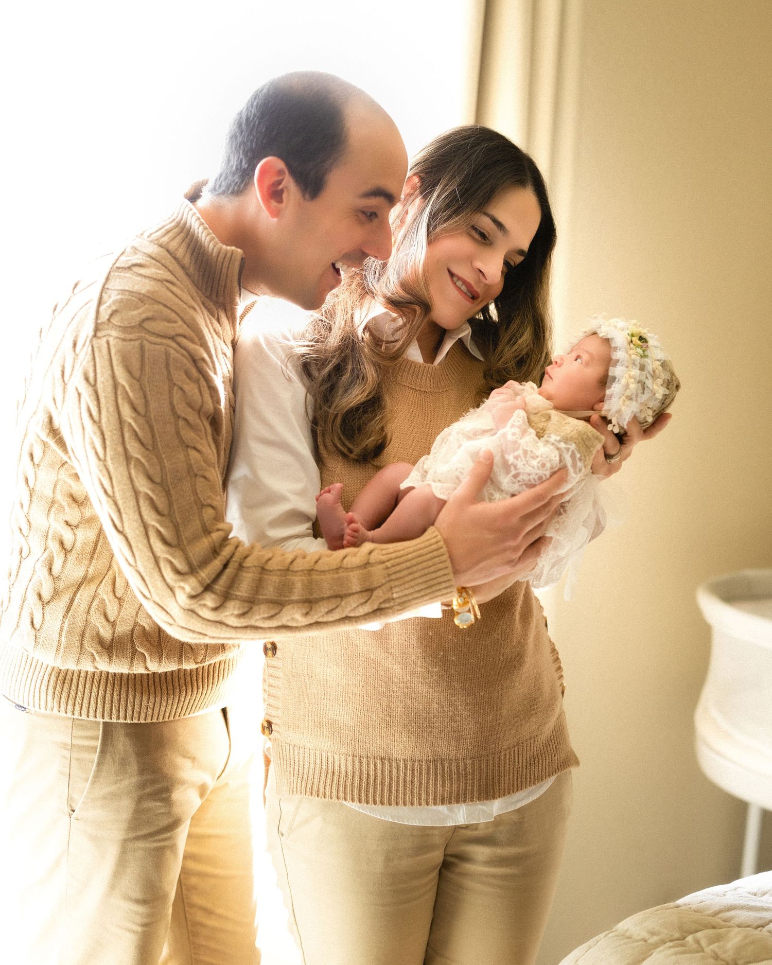 Happy parents in matching beige outfits lovingly admire their newborn baby girl wearing a floral bonnet near a bright window.