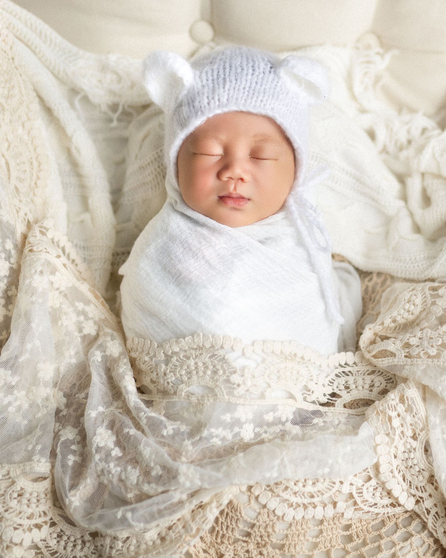 Sleeping newborn baby swaddled in white wrap wearing a knit bear ear bonnet on a lace blanket.