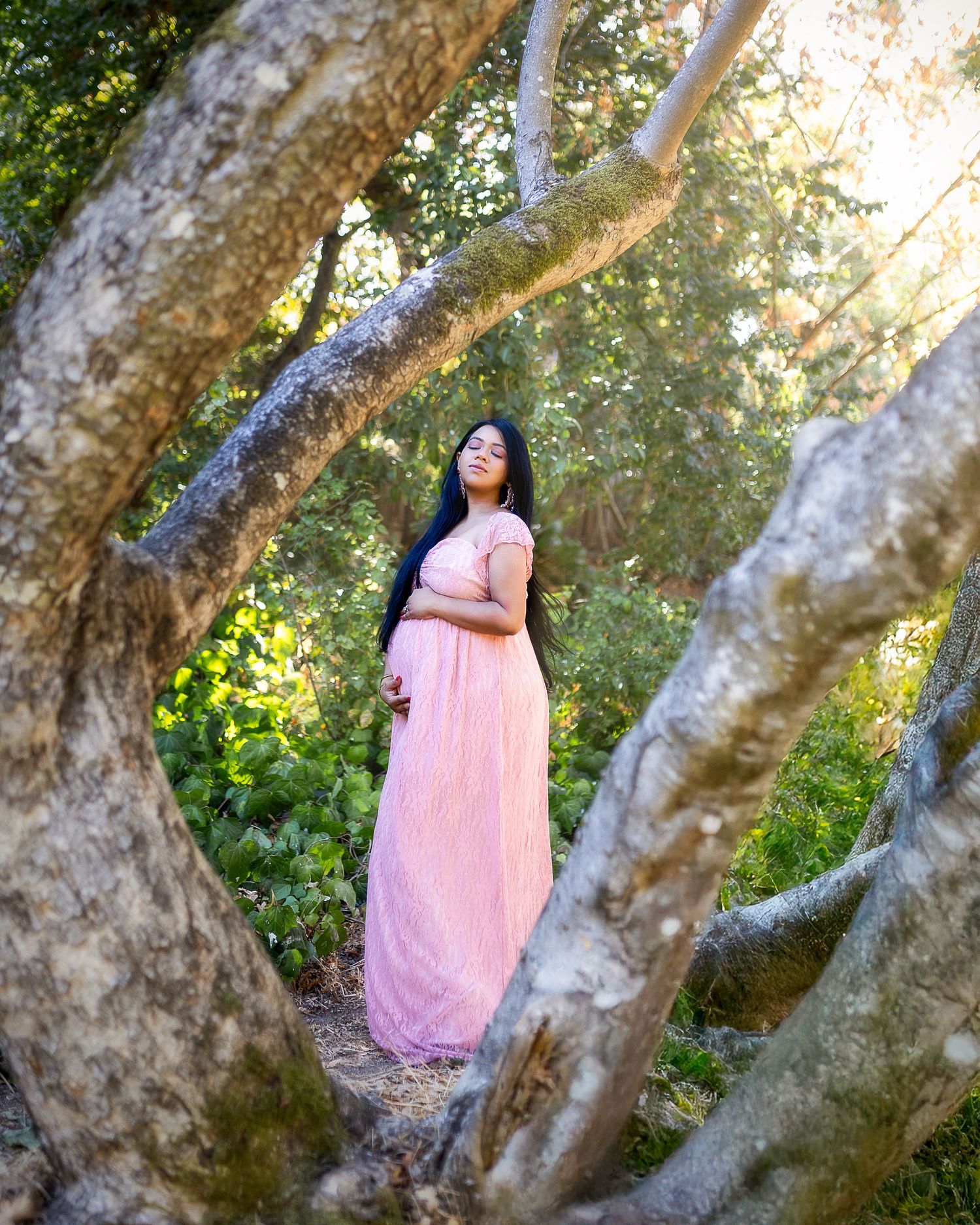 A person in a flowing pink dress poses gracefully among tree branches in a sunlit outdoor setting.