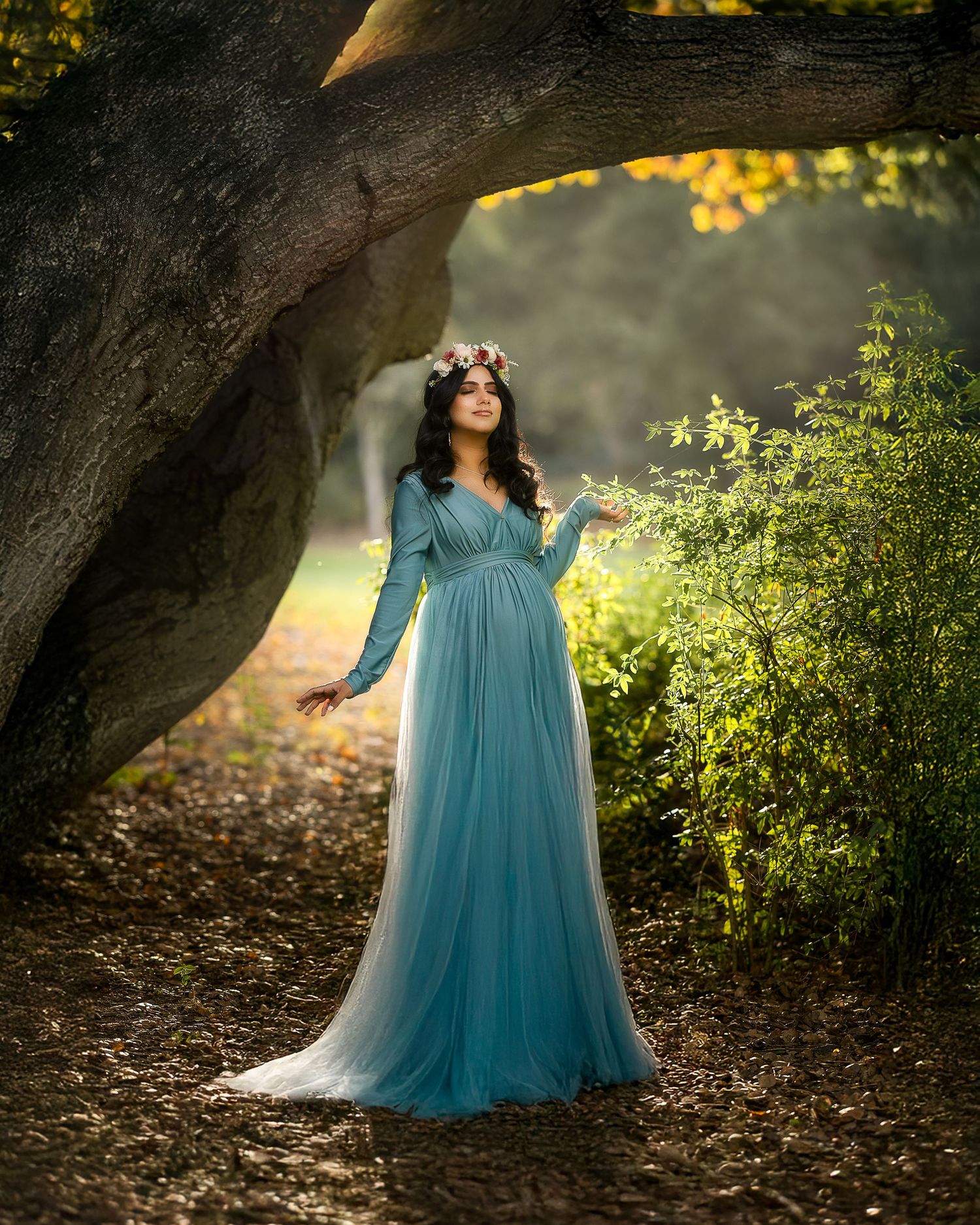 A woman in a flowing turquoise gown poses gracefully under a large tree in a dreamy outdoor setting.