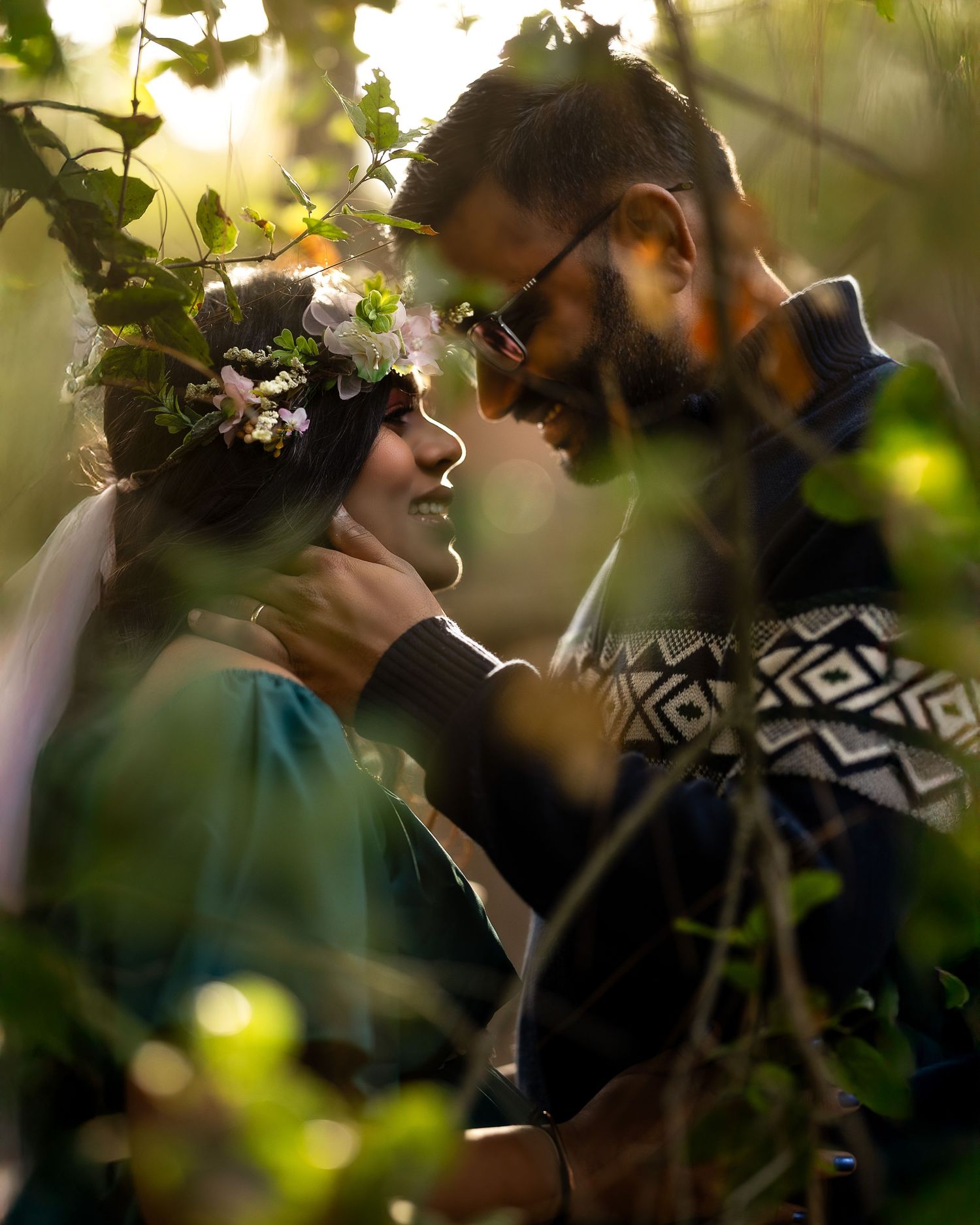Two people share an intimate moment outdoors with sunlight filtering through green foliage in the background.