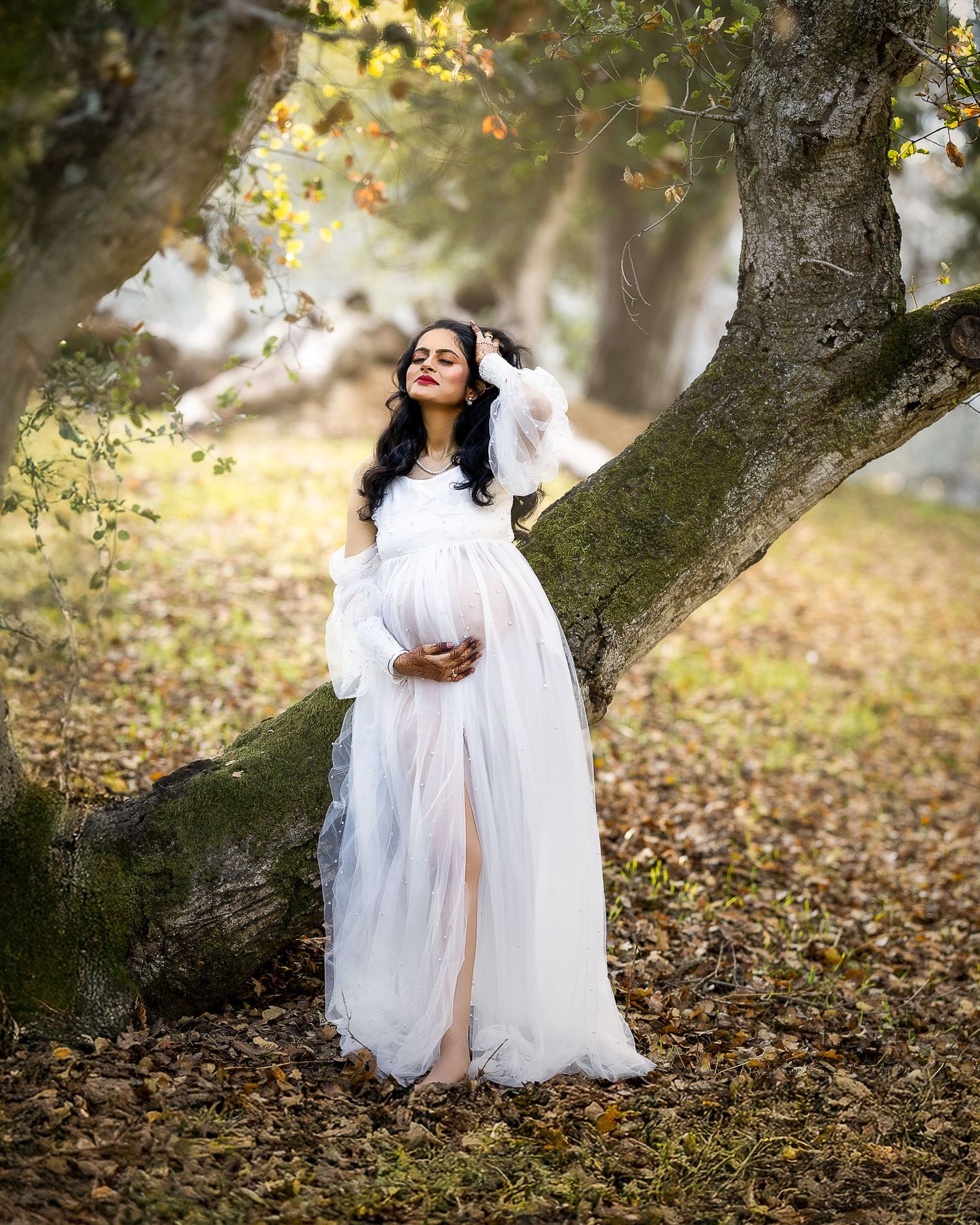A dreamy maternity photo in a white flowing dress under autumn trees with soft natural lighting and fallen leaves.