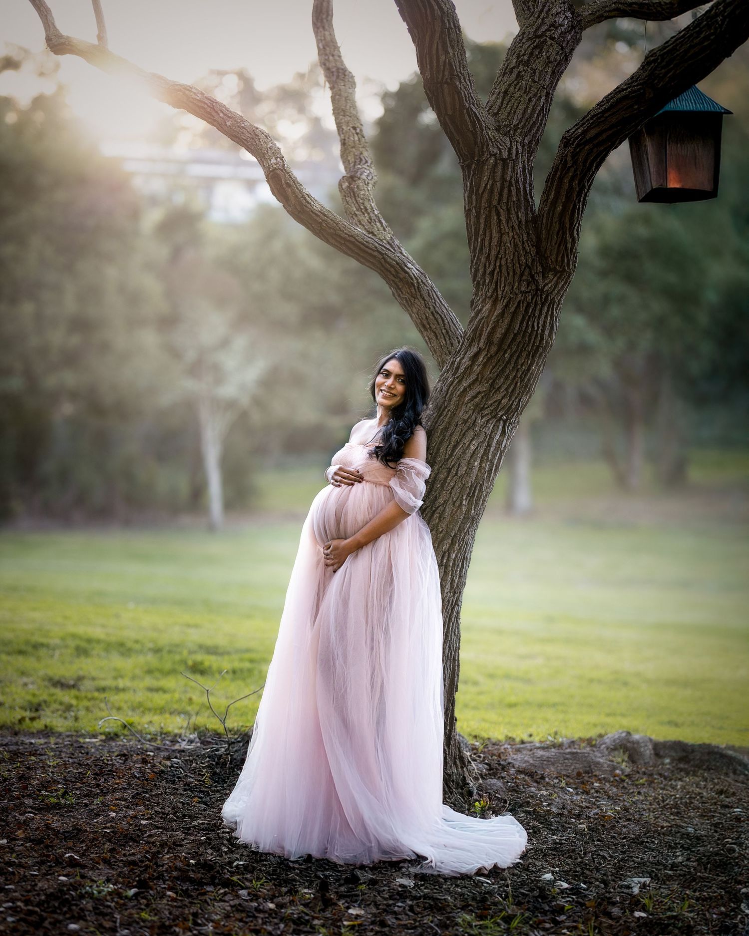 A person in a flowing pink tulle gown poses gracefully against a large tree trunk in a dreamy outdoor setting at sunset.