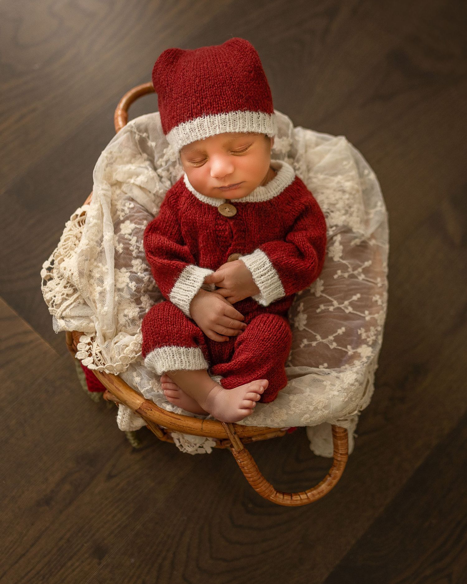 Sleeping baby in red and white knit outfit rests peacefully in vintage wicker bassinet with lace bedding.