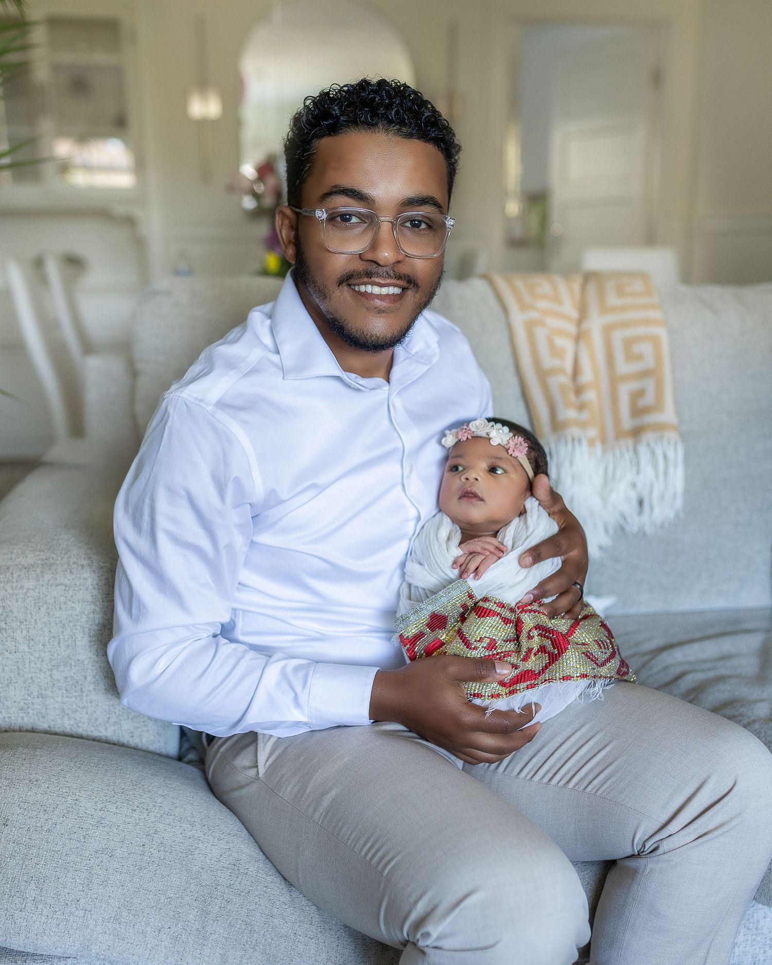 A parent wearing a white shirt and grey pants sits on a couch while holding and bonding with a small baby.