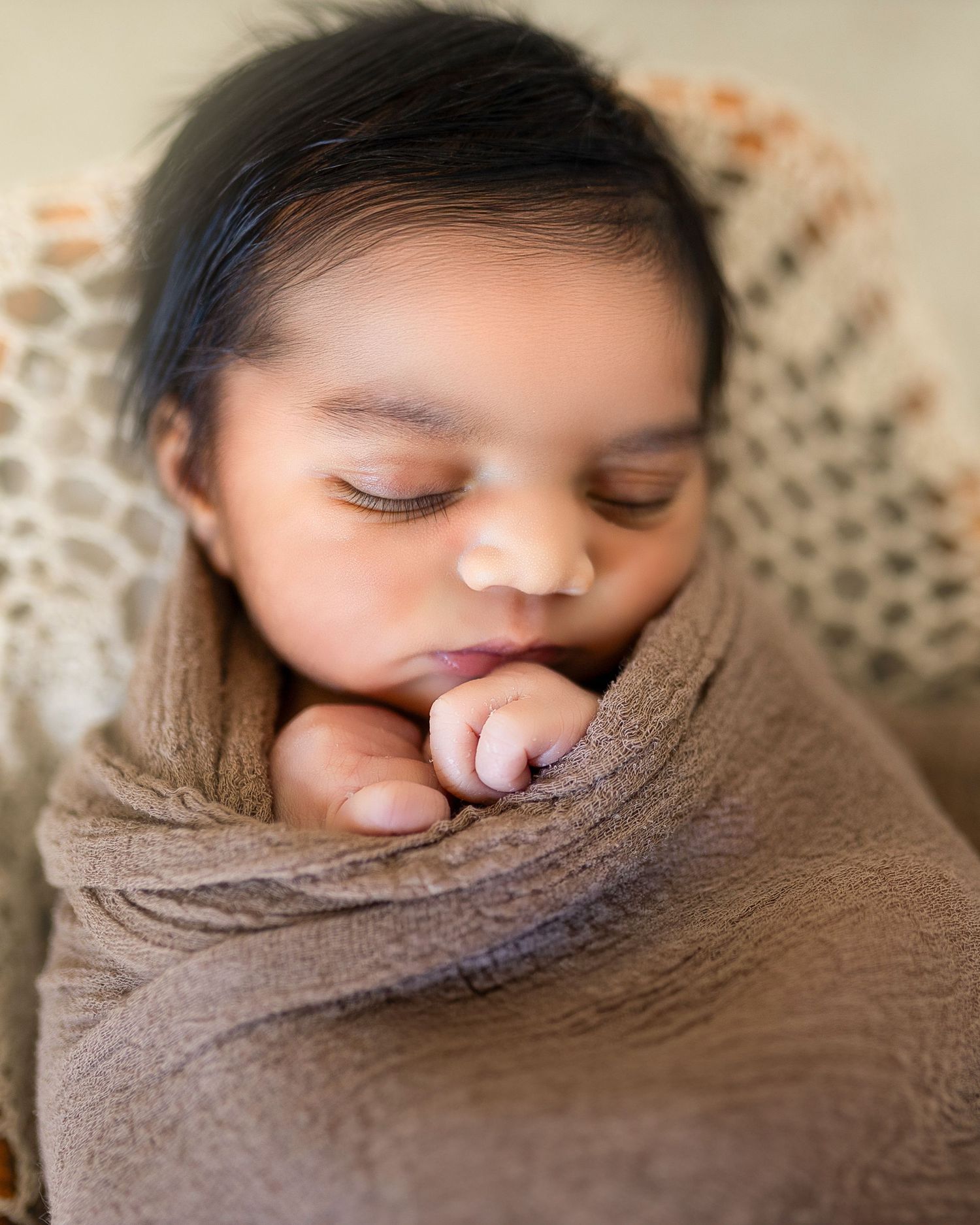 A sleeping newborn wrapped in a brown swaddle blanket with soft lighting creating a peaceful and serene portrait.