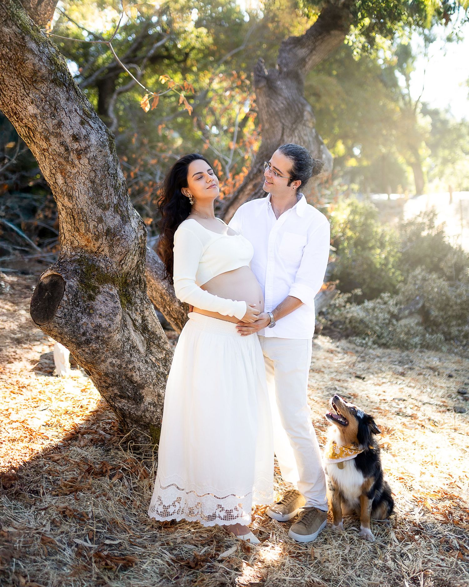 Romantic couple in white outfits share tender moments under a tree during golden hour sunset photoshoot with their dog.