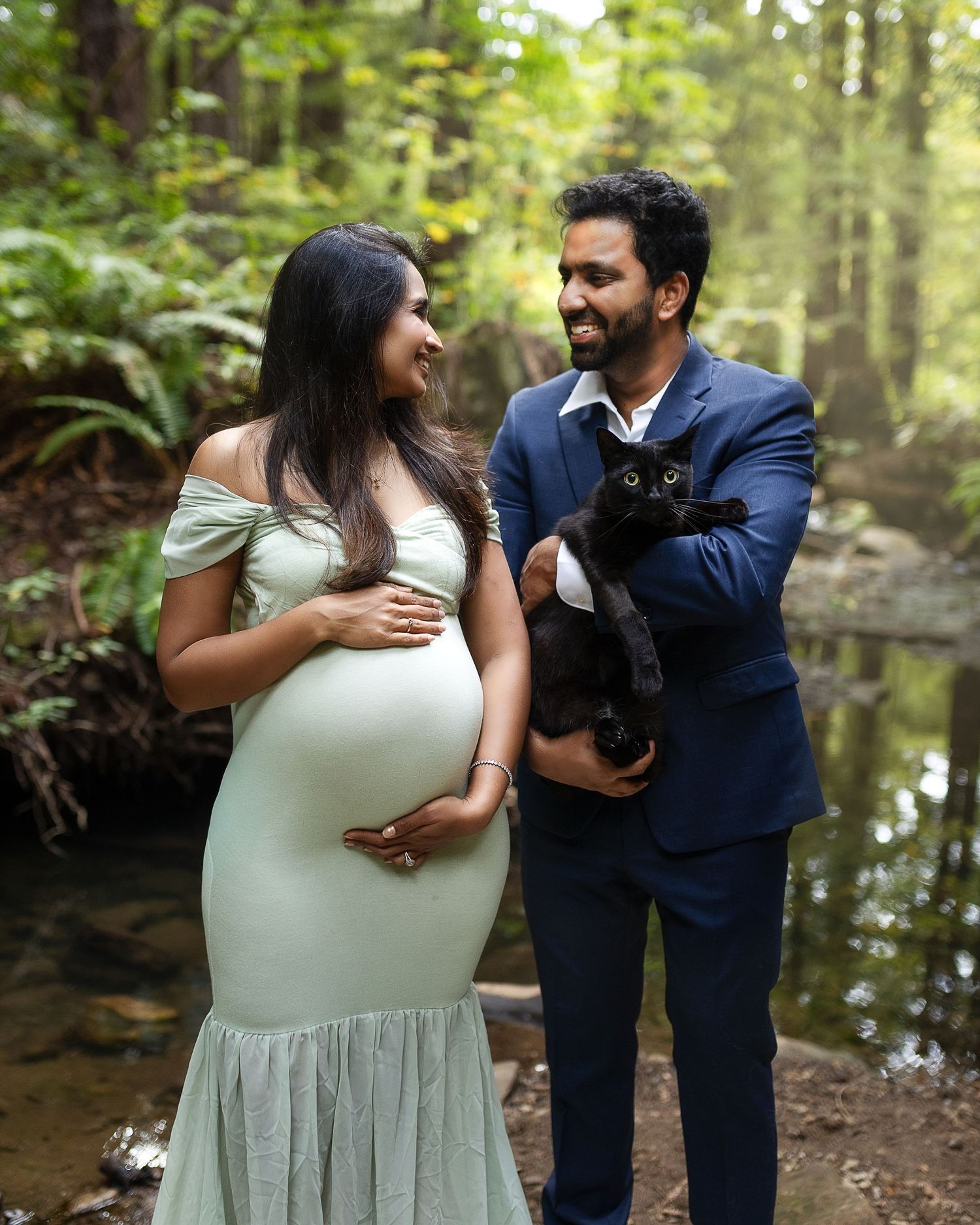 A couple in a wooded setting poses together with their black dog for a maternity photoshoot near a stream.
