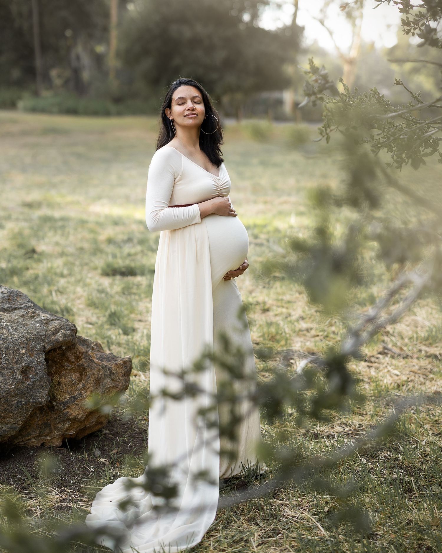 A person in a flowing white dress poses gracefully in a sunlit outdoor setting with natural foliage and trees.
