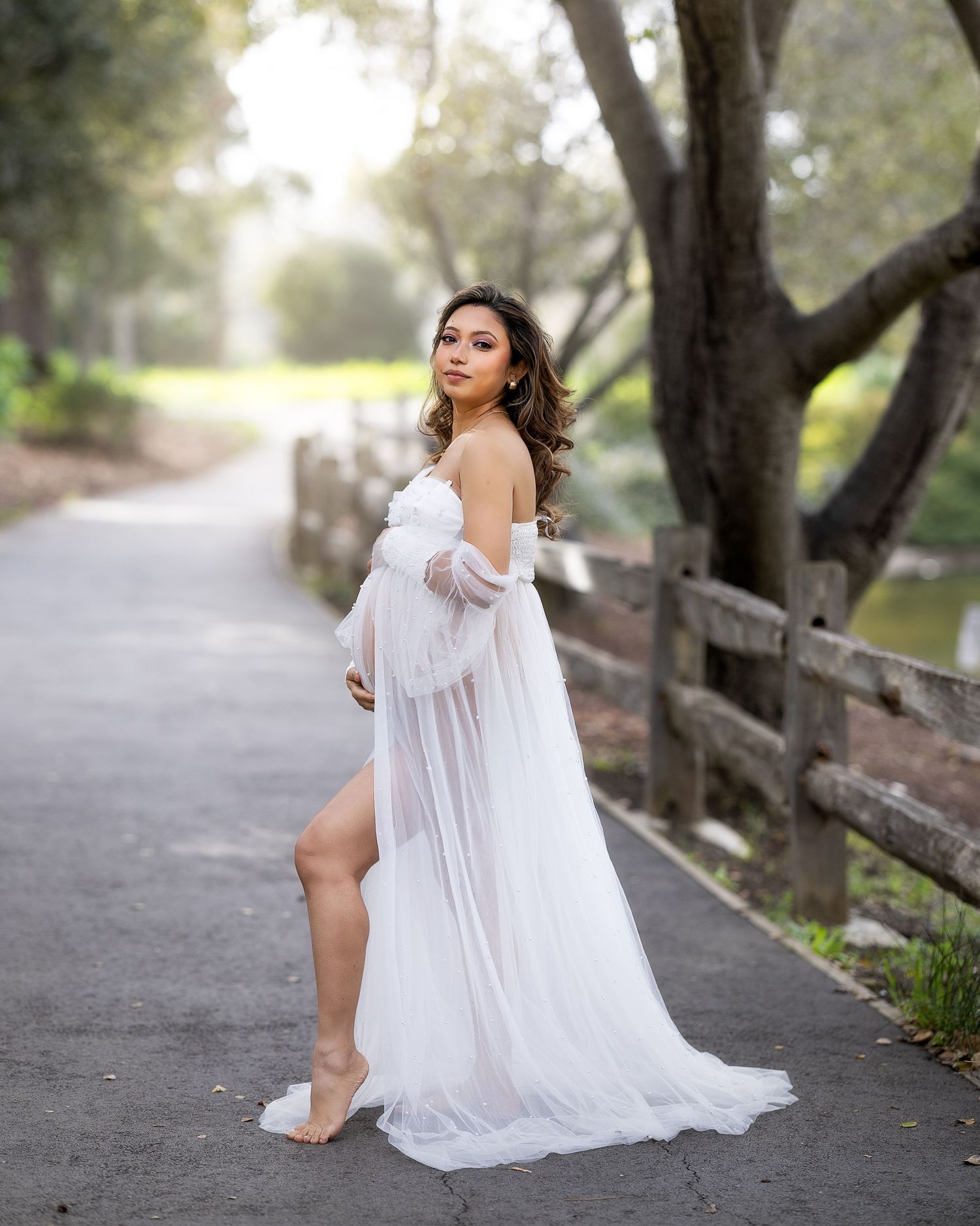 A flowing white dress creates an ethereal silhouette against a misty tree-lined path during an outdoor photo session.