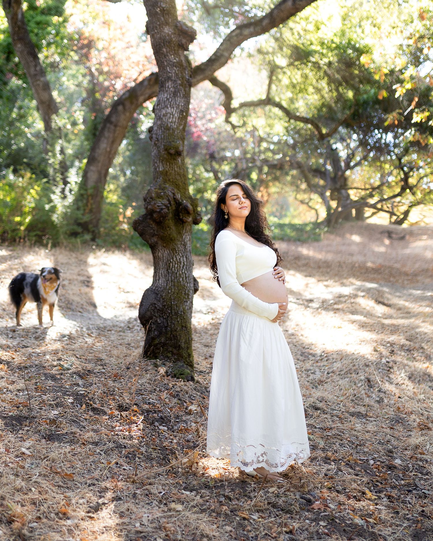 A person in a flowing white dress poses gracefully among oak trees in a sun-dappled forest setting.