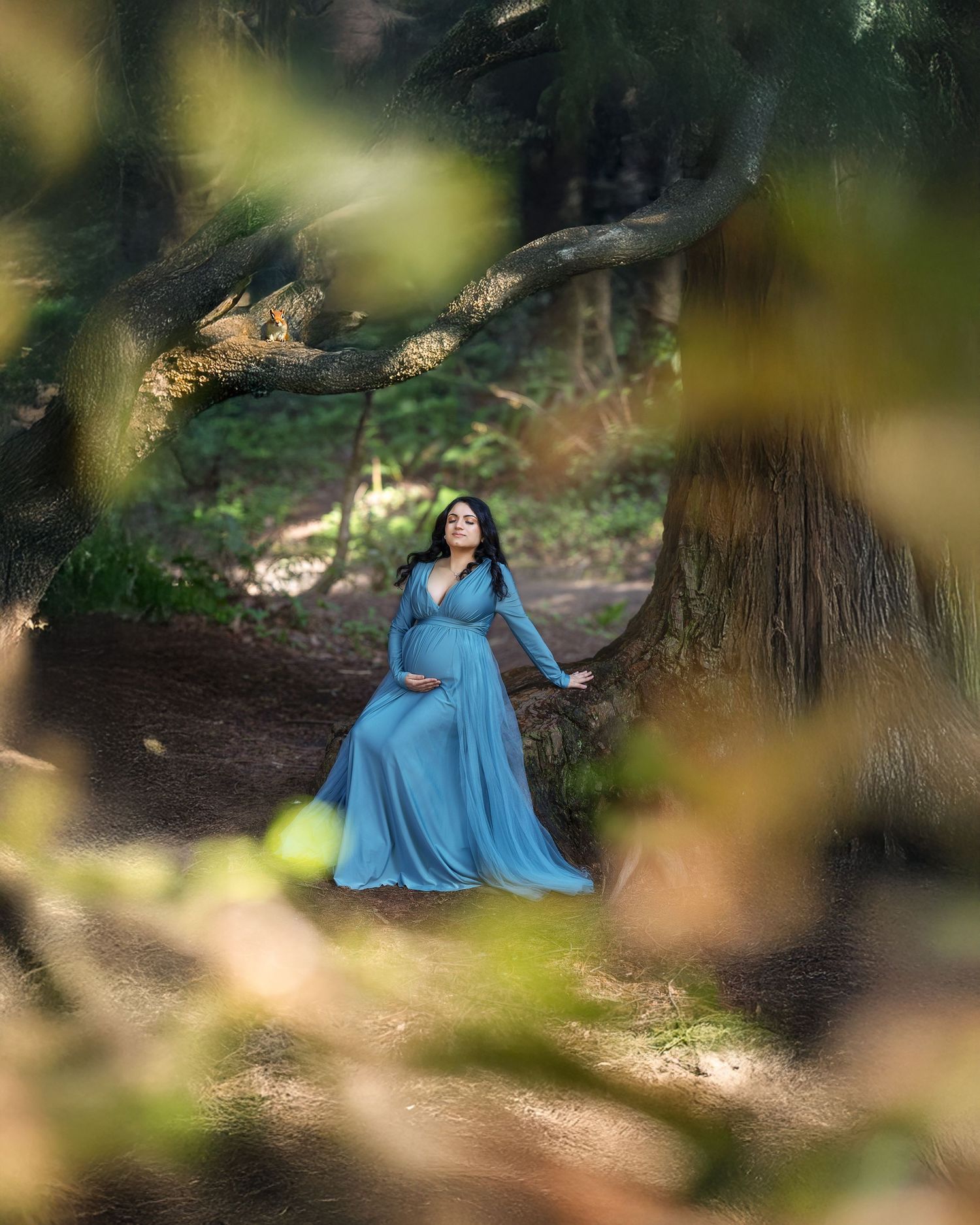 A person in a flowing blue dress poses gracefully in a dreamy forest setting with dappled sunlight.