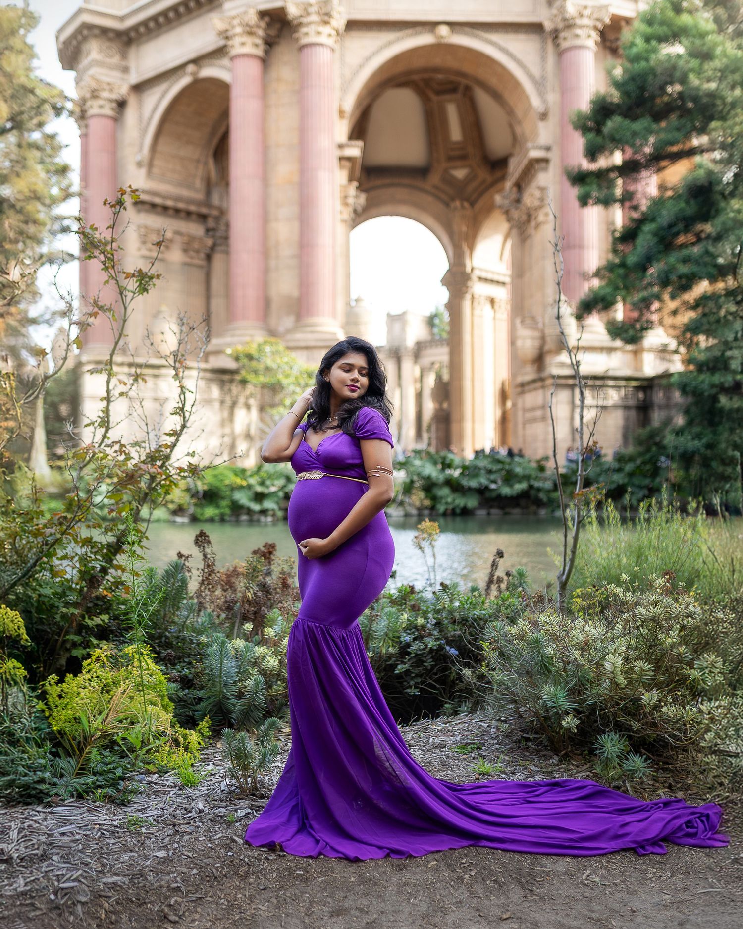 A woman in a flowing purple maternity gown poses gracefully in a garden setting with classical architecture.
