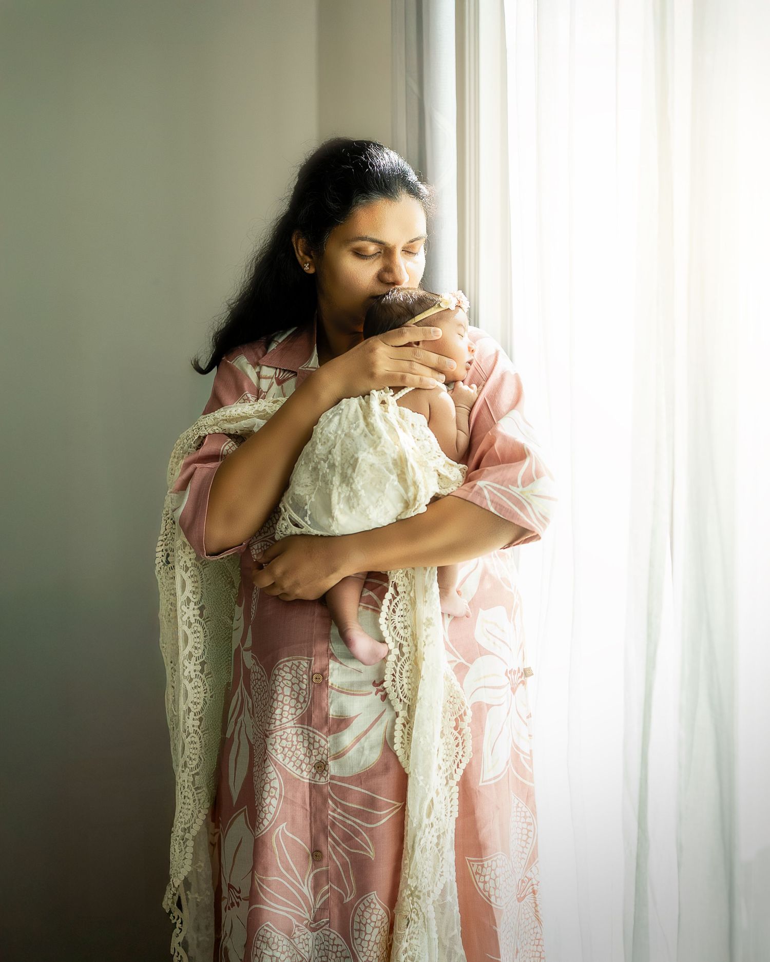 A tender moment by a window as a mother in a pink floral dress cradles and kisses her newborn wrapped in lace.
