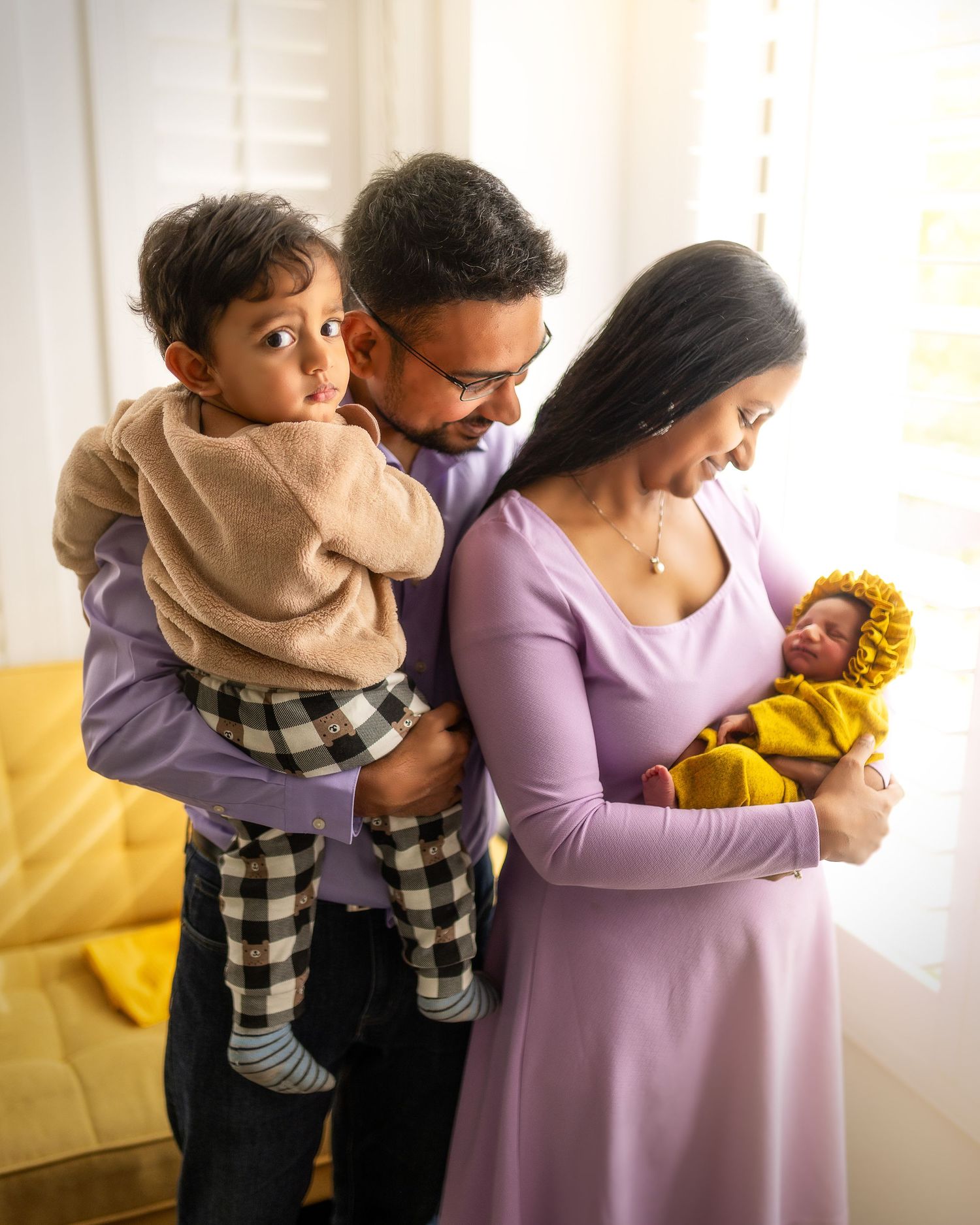 A family shares a tender moment together on a yellow couch near a bright window.