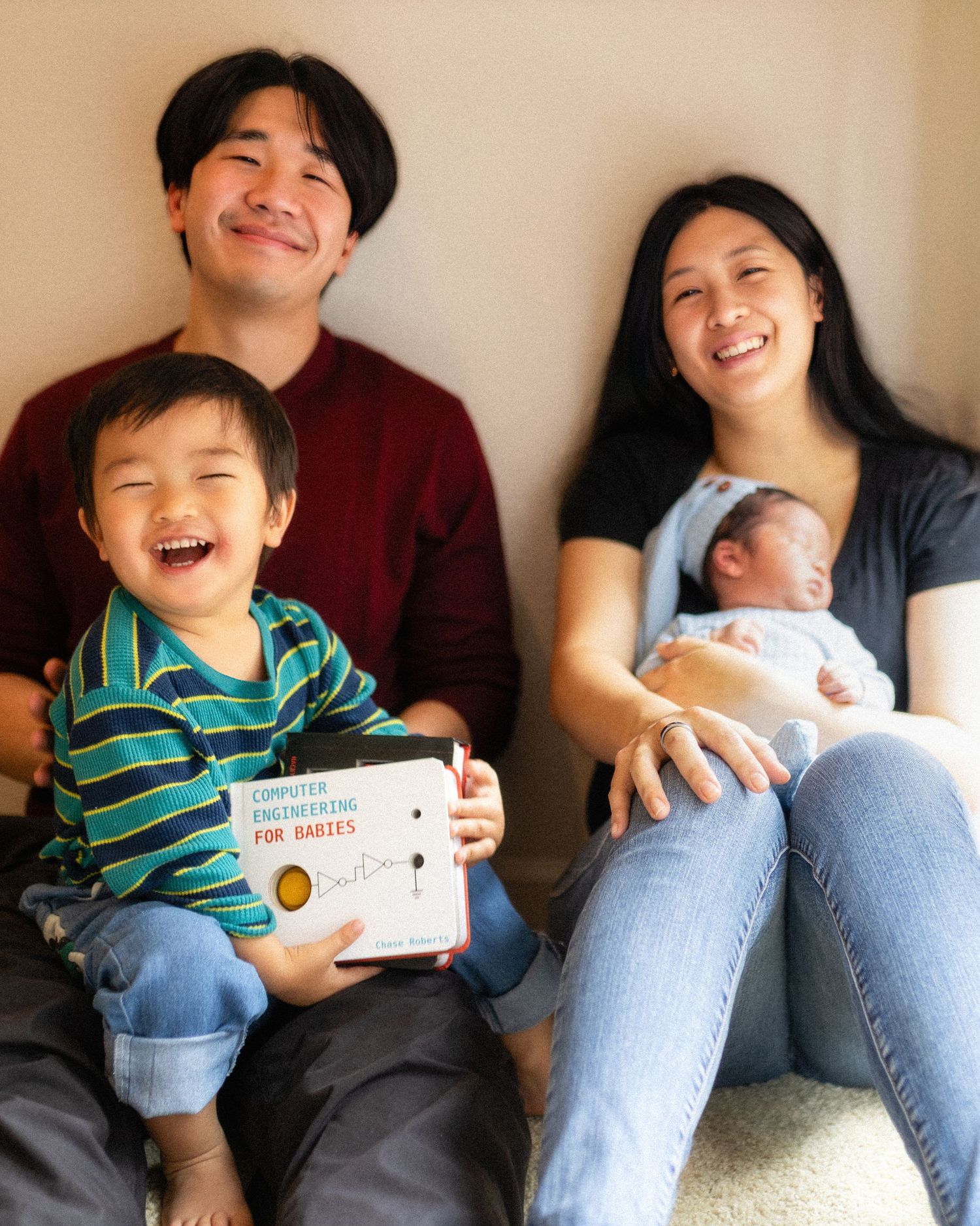 Happy Asian family of four sitting together, parents smiling with toddler holding a Computer Engineering for Babies book and newborn.