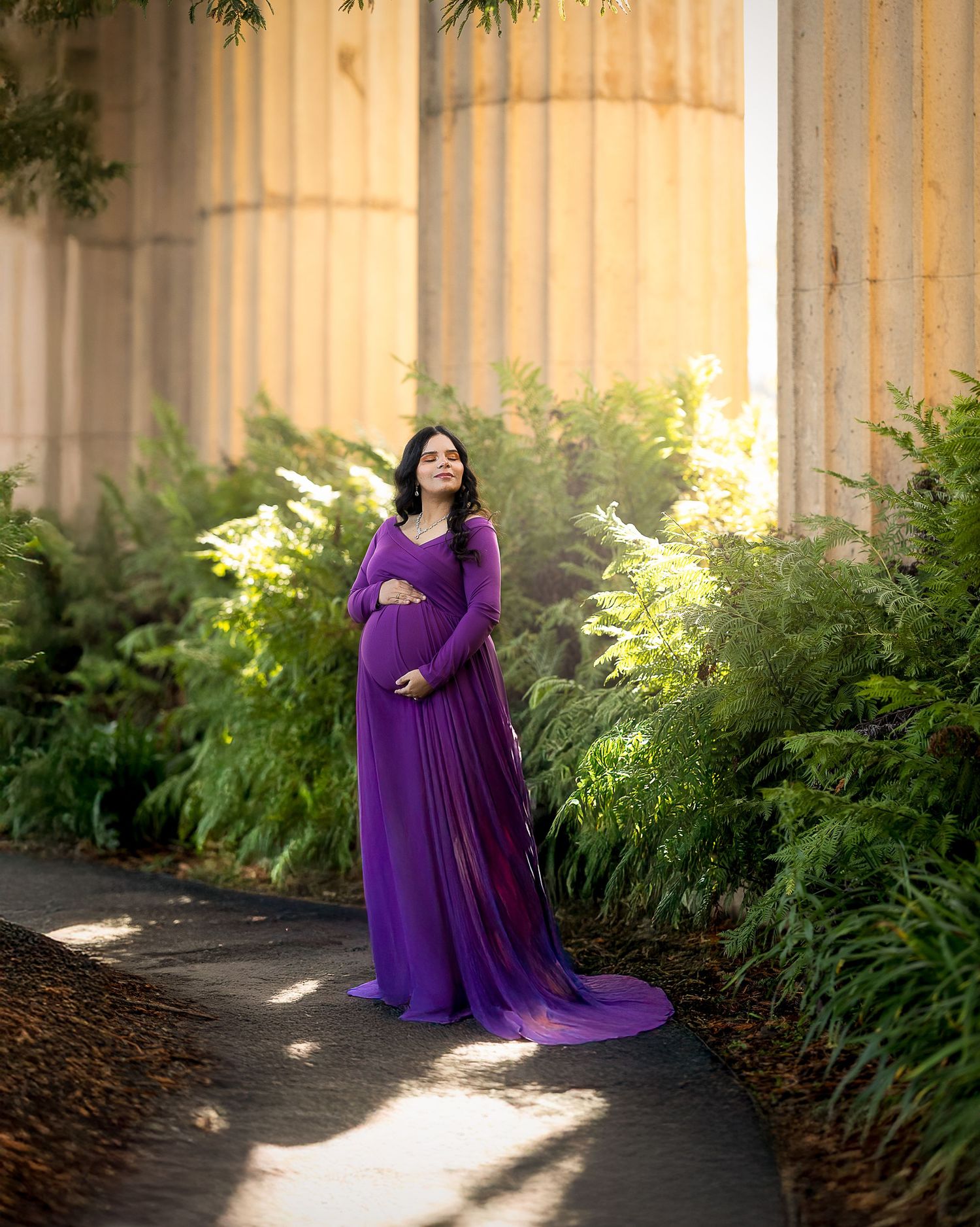 Expectant mother in flowing purple maternity gown poses elegantly among ferns and columns in soft natural lighting.