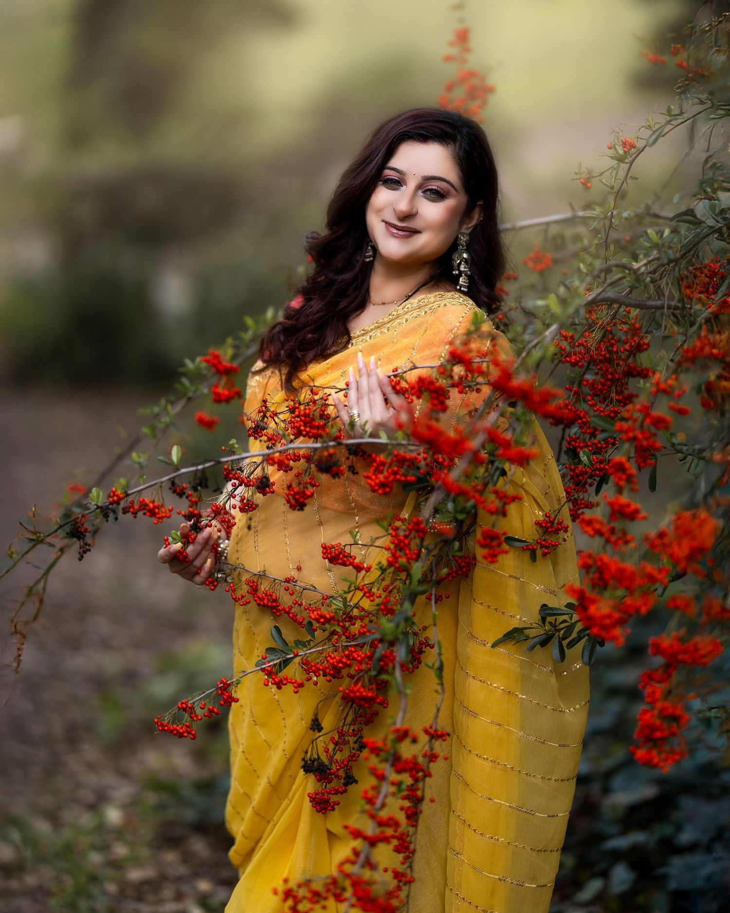 A woman in a vibrant yellow sari poses elegantly among red berries and foliage in a garden setting.