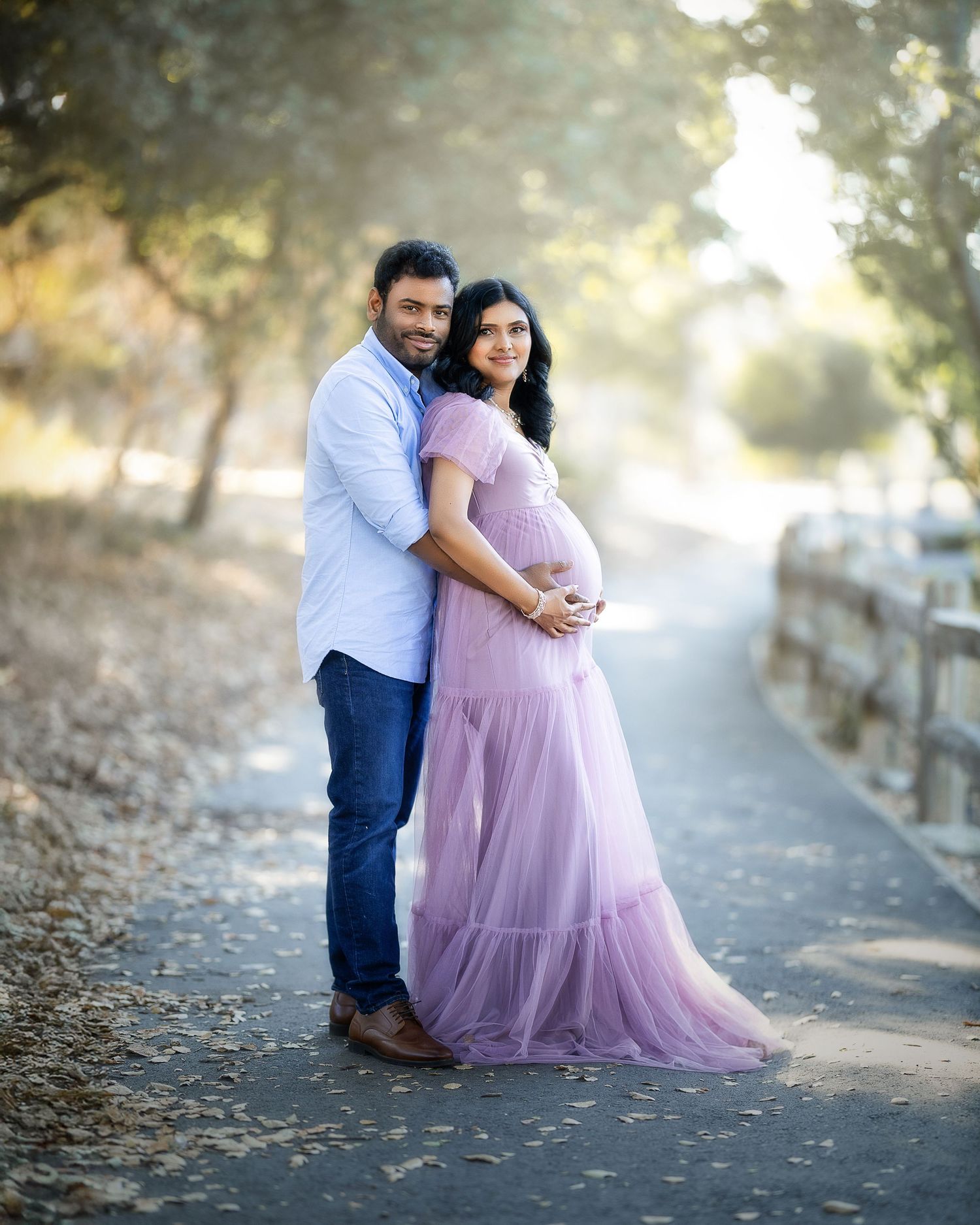 A romantic maternity photoshoot in a park with soft sunlight filtering through trees and a flowing lavender dress.