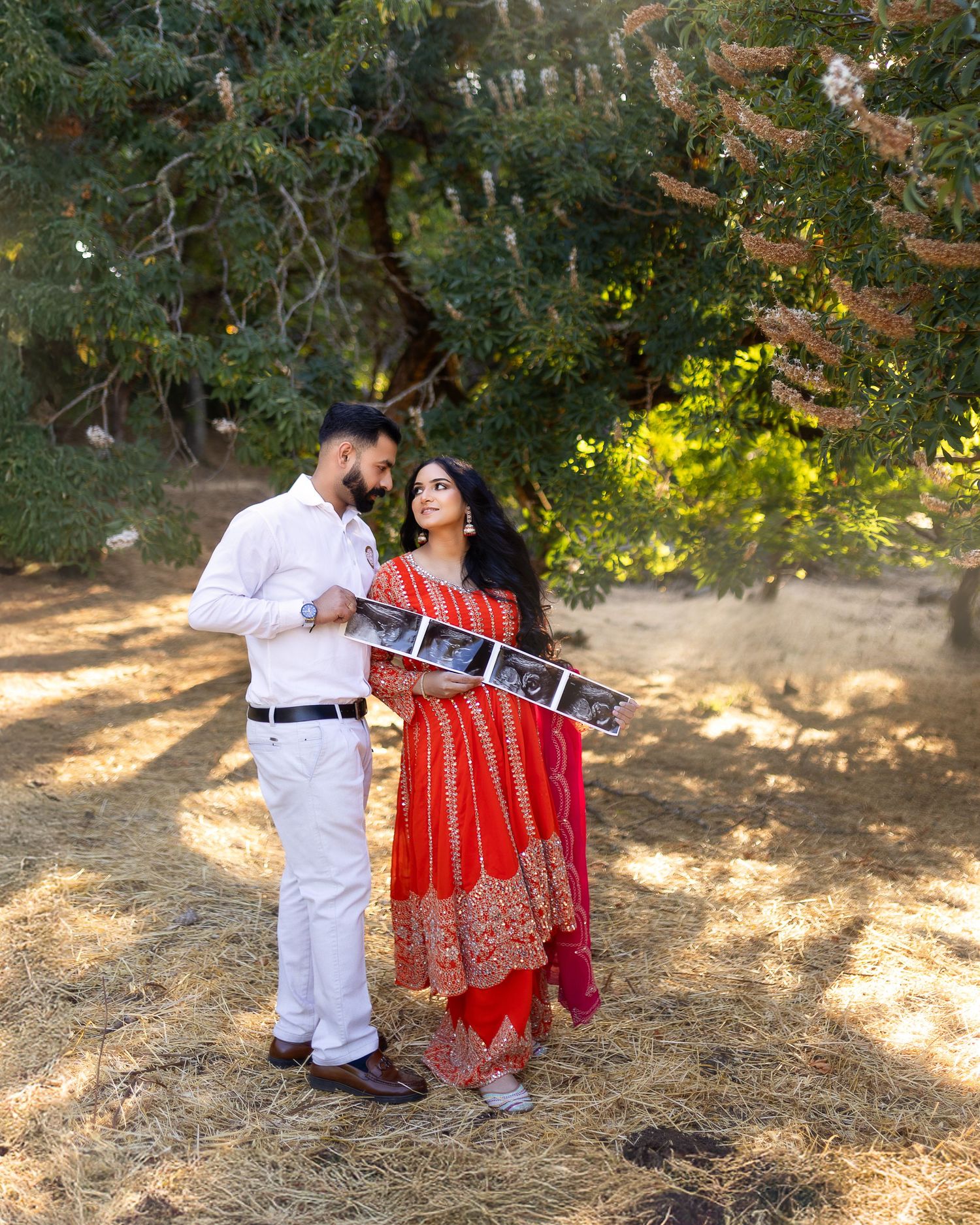A couple in traditional Indian attire poses together outdoors under trees in a romantic photoshoot setting.