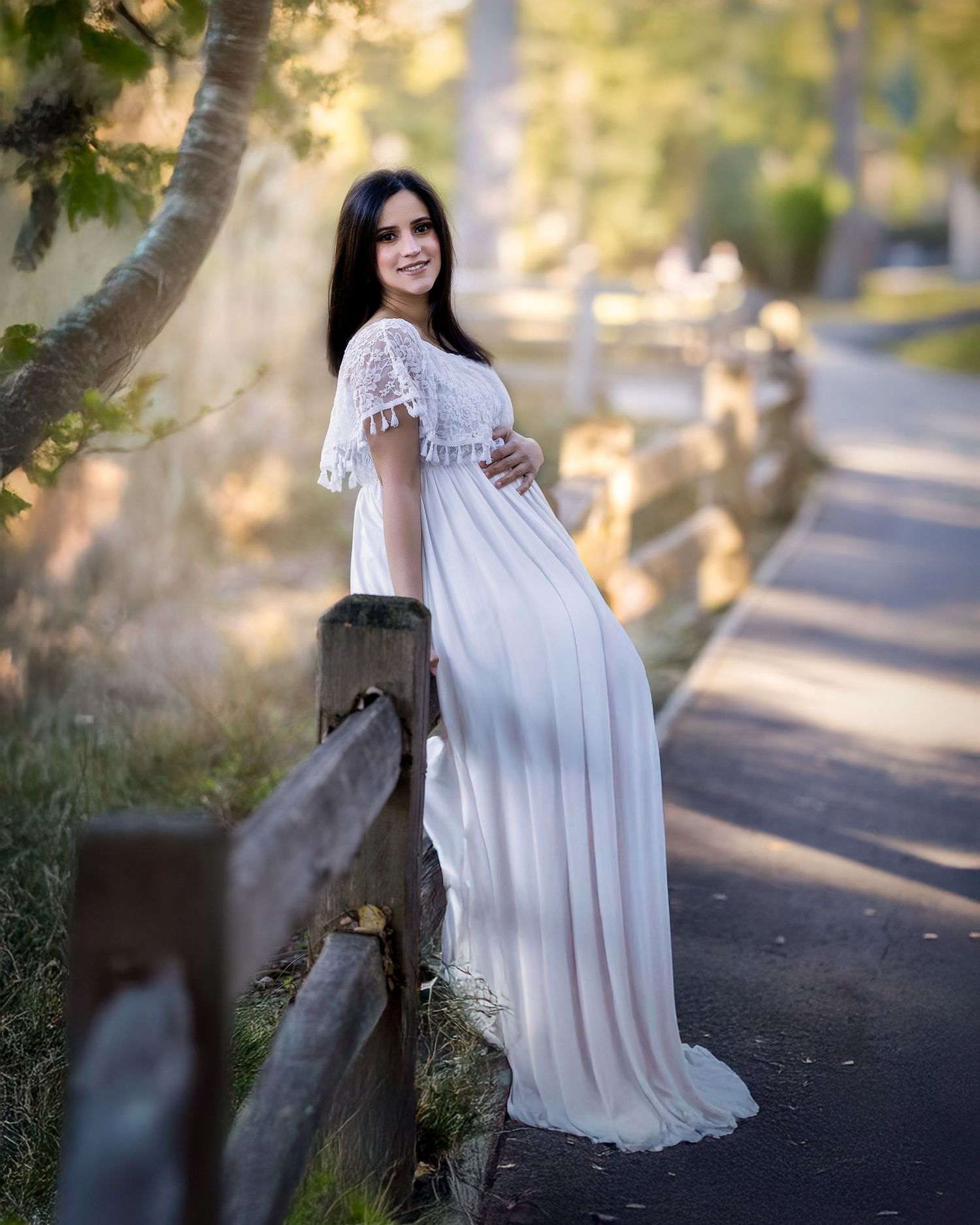 A flowing white dress creates elegant movement against a sunlit outdoor path with trees in the background.