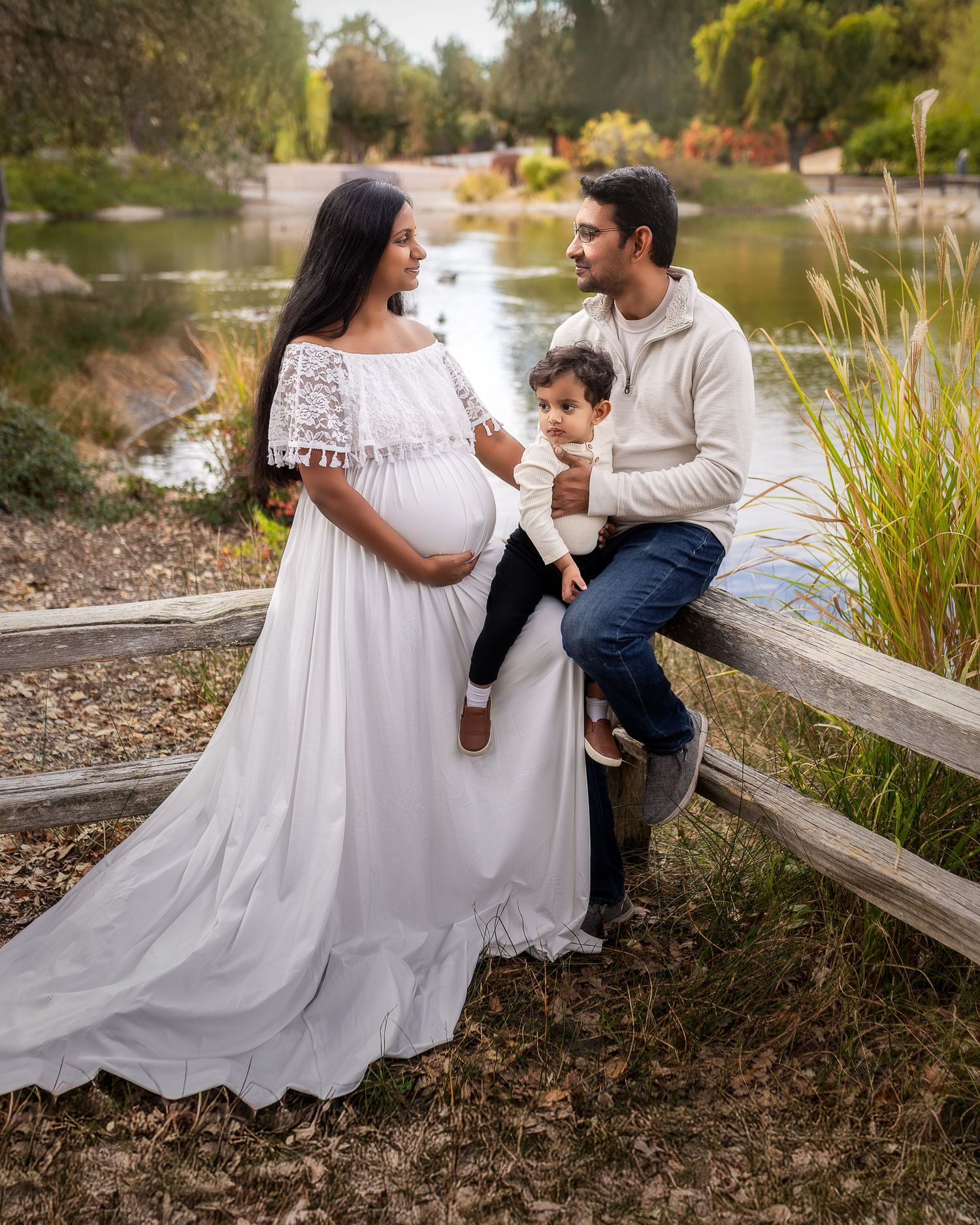 A couple shares an intimate moment outdoors near a lake with tall grasses and a wooden fence in the background.