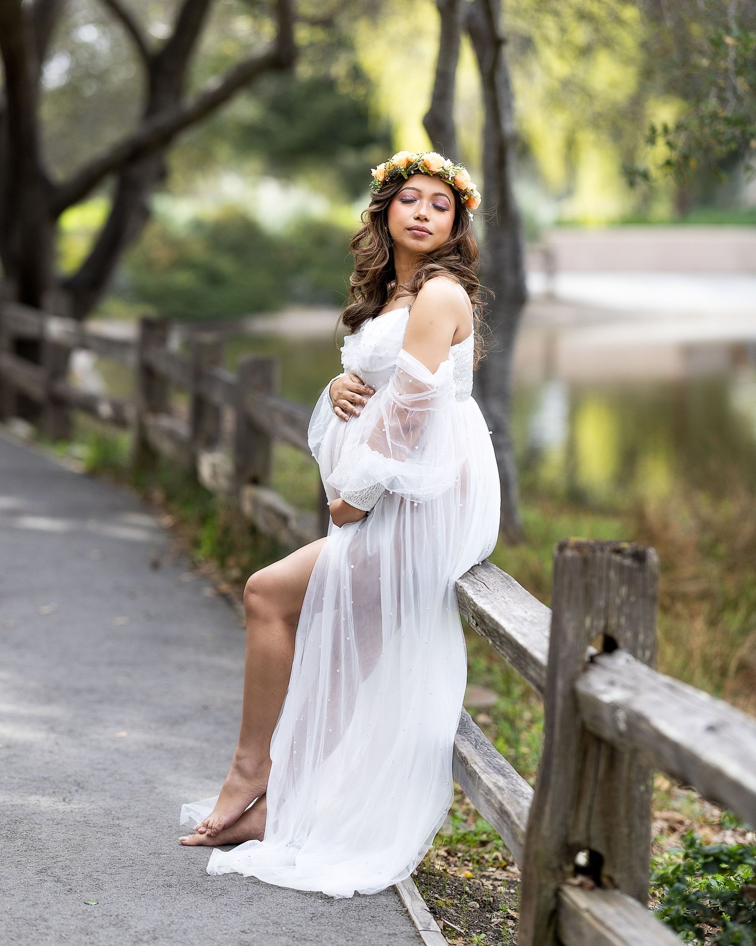 Ethereal outdoor portrait in flowing white dress sitting on wooden fence in dappled sunlight.