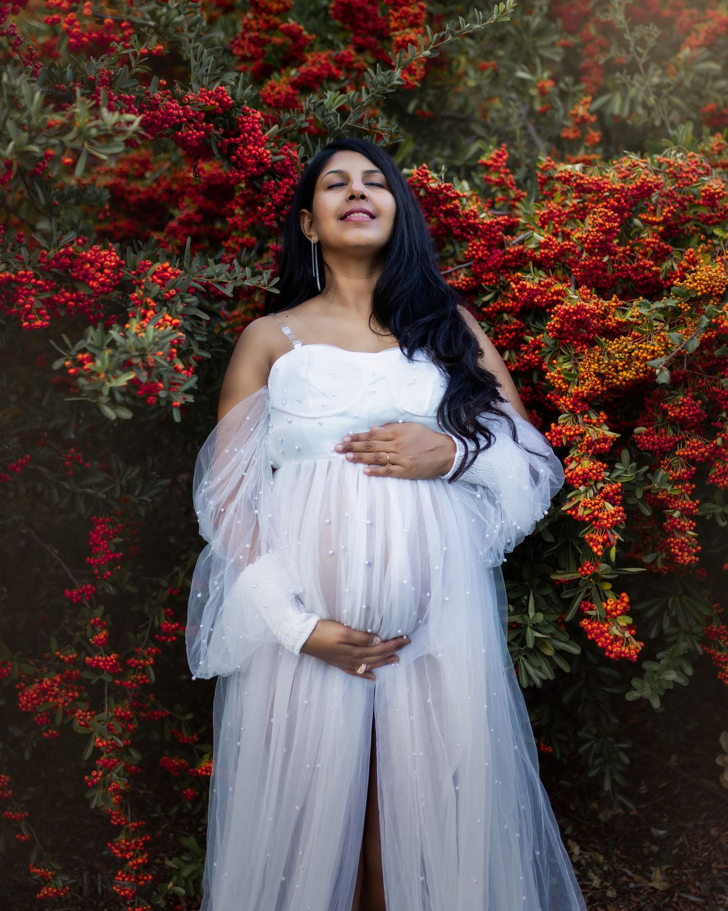 A flowing white dress contrasts beautifully against vibrant red berries and foliage in an outdoor portrait setting.