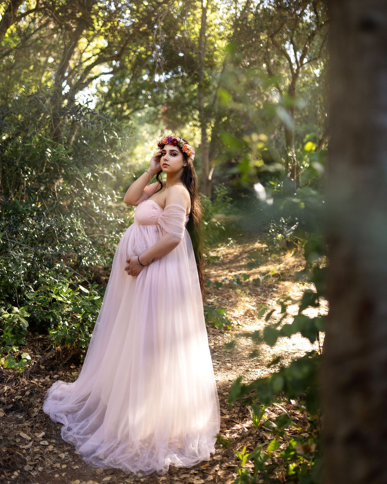 Ethereal figure in a flowing pink gown stands gracefully among sunlit trees in an enchanted forest setting.