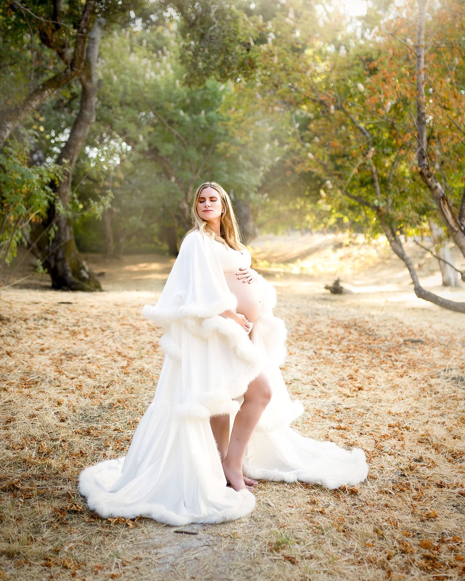 A person in a flowing white dress sits gracefully on fallen autumn leaves in a sunlit outdoor setting.