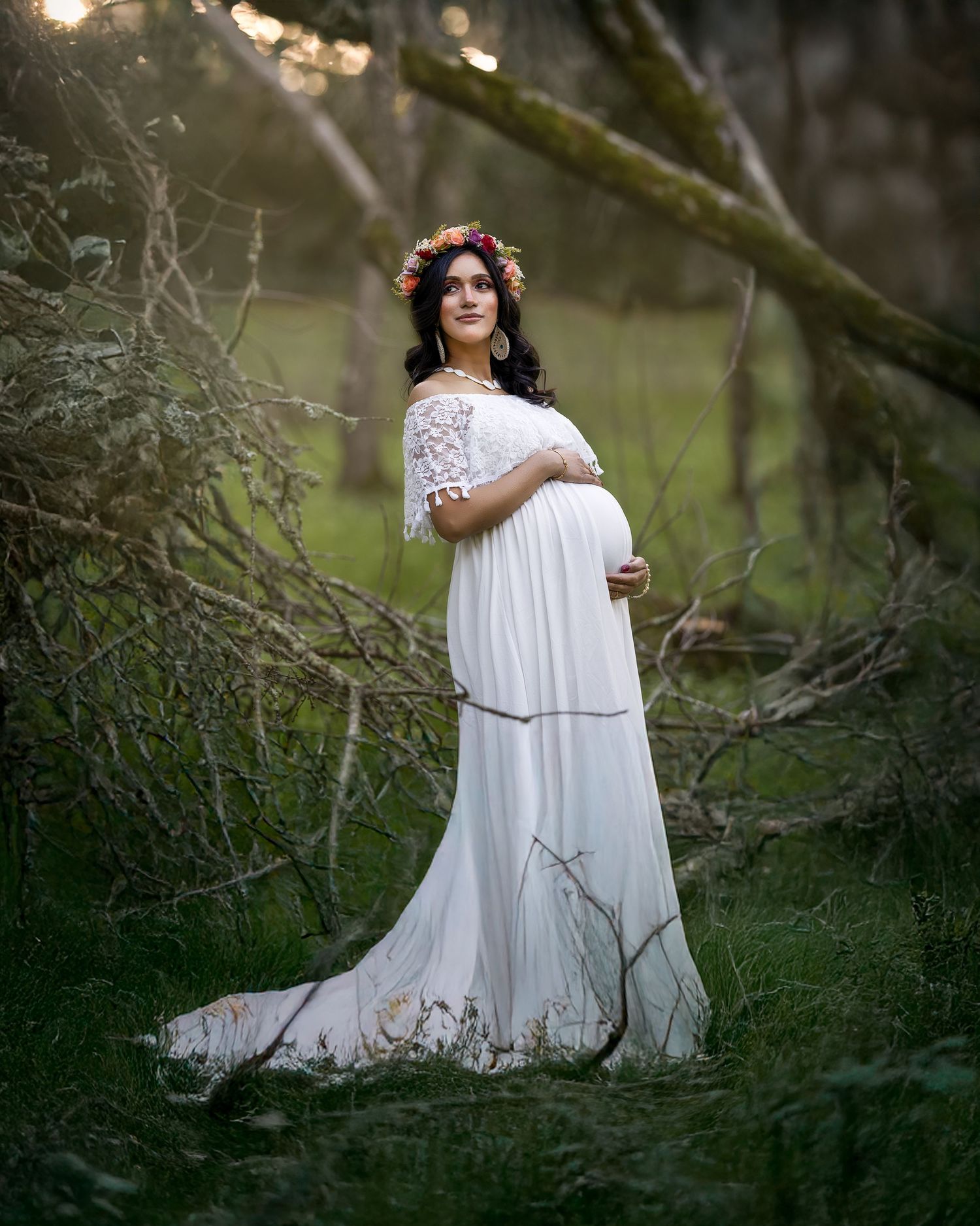 Ethereal outdoor portrait in white flowing dress with floral crown amid natural woodland setting at sunset.