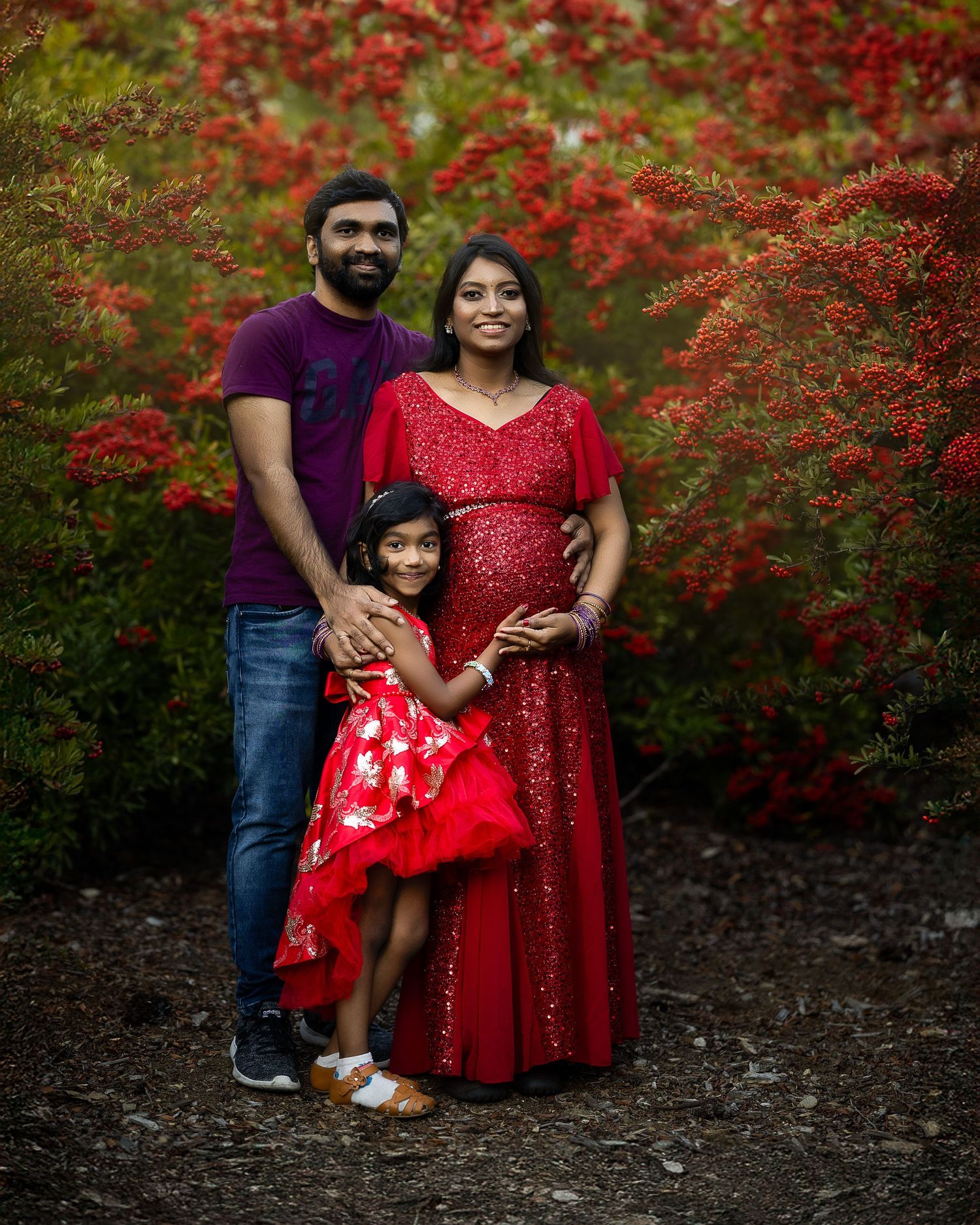 Family wearing coordinated red outfits poses together against vibrant red flowering bushes in an outdoor setting.
