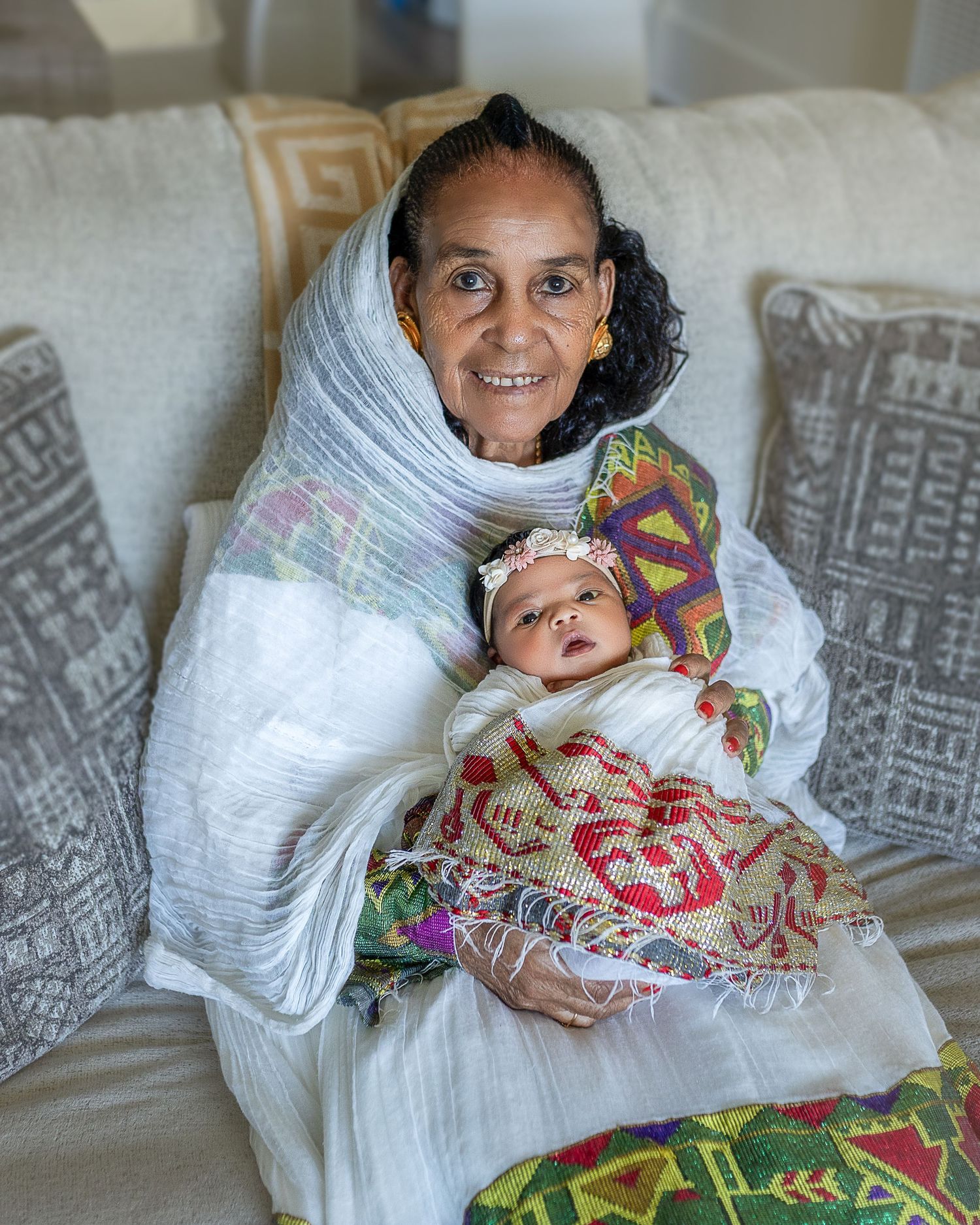 A figure in a white shawl sits on a couch holding a small baby wrapped in colorful patterned fabrics.