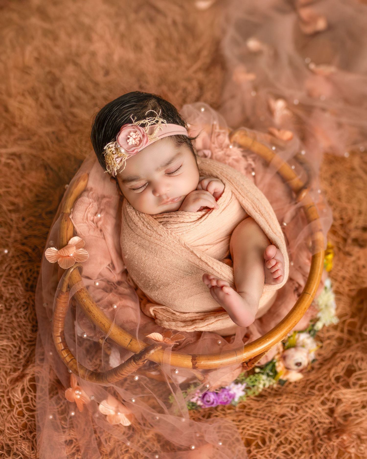 A sleeping newborn wrapped in peach fabric lies in a wooden bowl surrounded by delicate flowers and soft lighting.