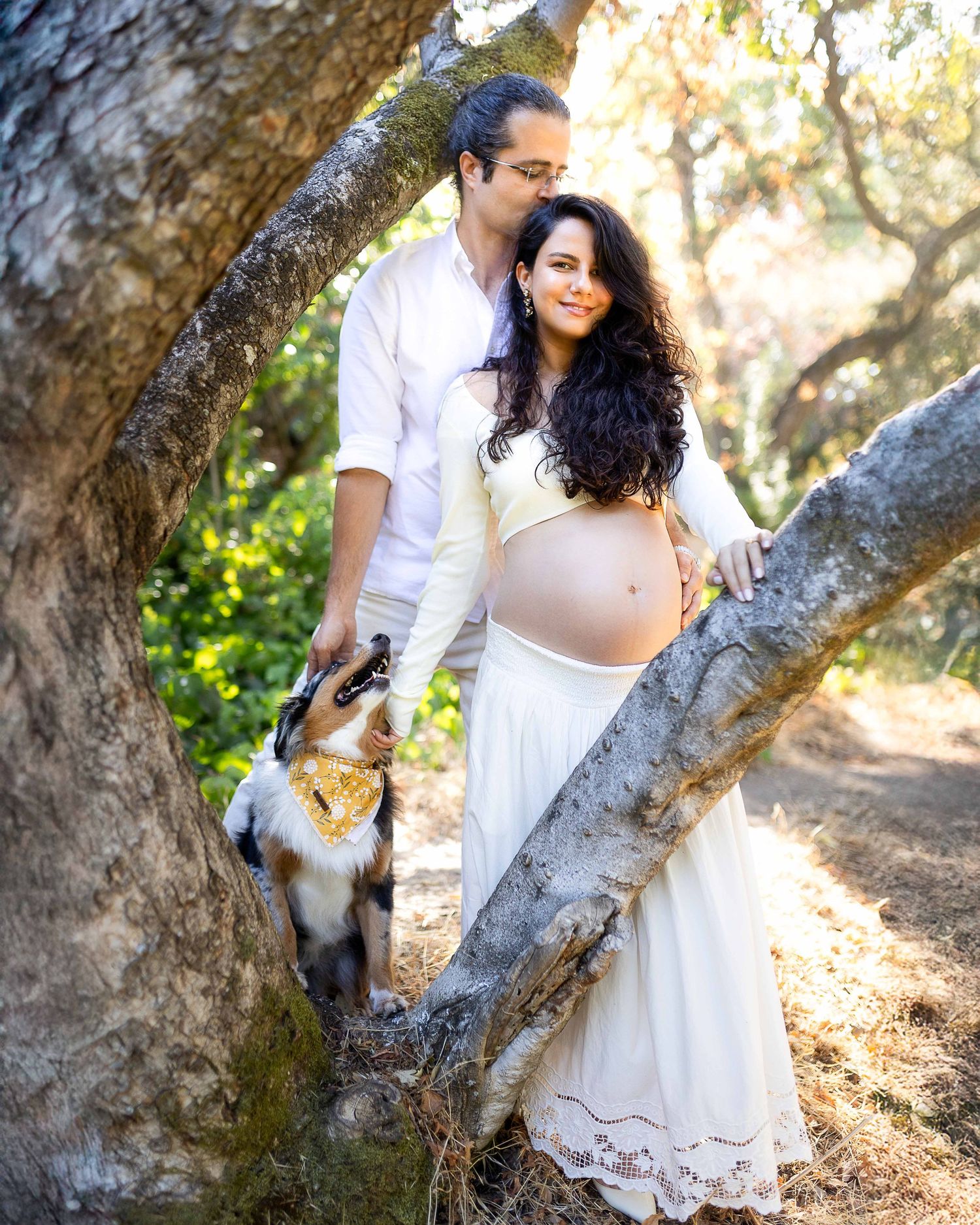 Couple in white outfits poses with dog among trees during outdoor maternity photoshoot in sunlit forest.