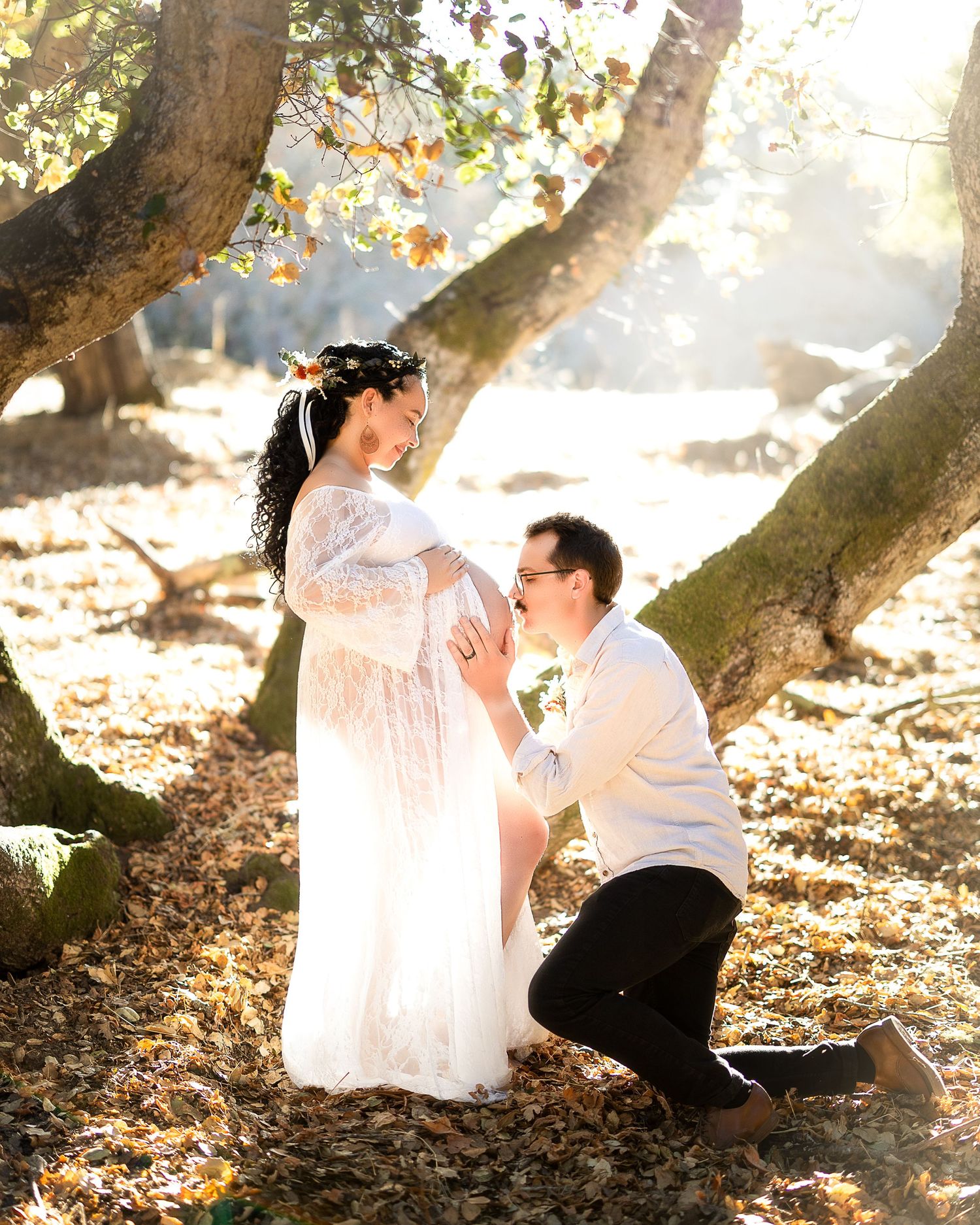 A romantic maternity photo shows a tender moment between a couple under sunlit trees with autumn leaves.