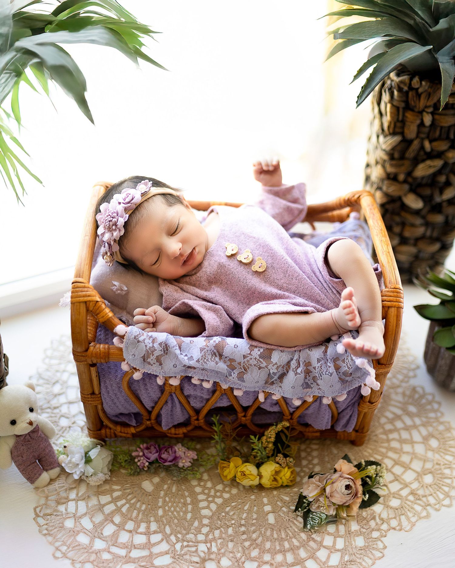 A baby in a purple dress sleeps peacefully in a rustic wooden basket surrounded by flowers and a lace doily.