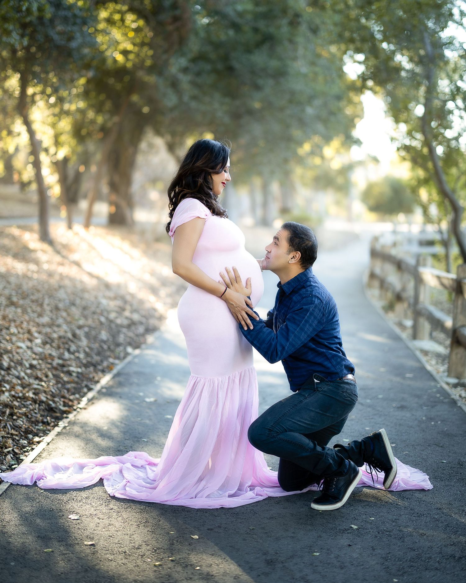 A tender maternity moment captured on a tree-lined path with soft sunlight filtering through the leaves.