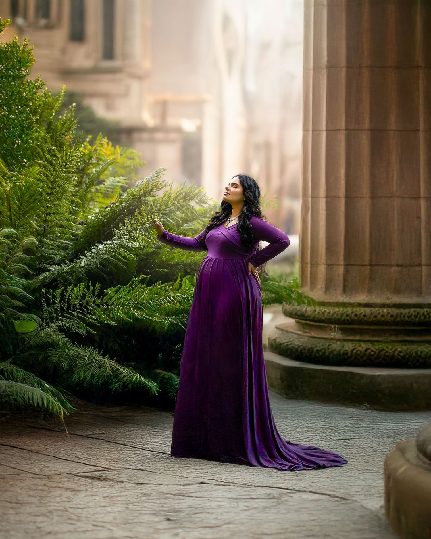 A flowing purple gown creates an elegant silhouette against stone columns and lush ferns in a garden setting.