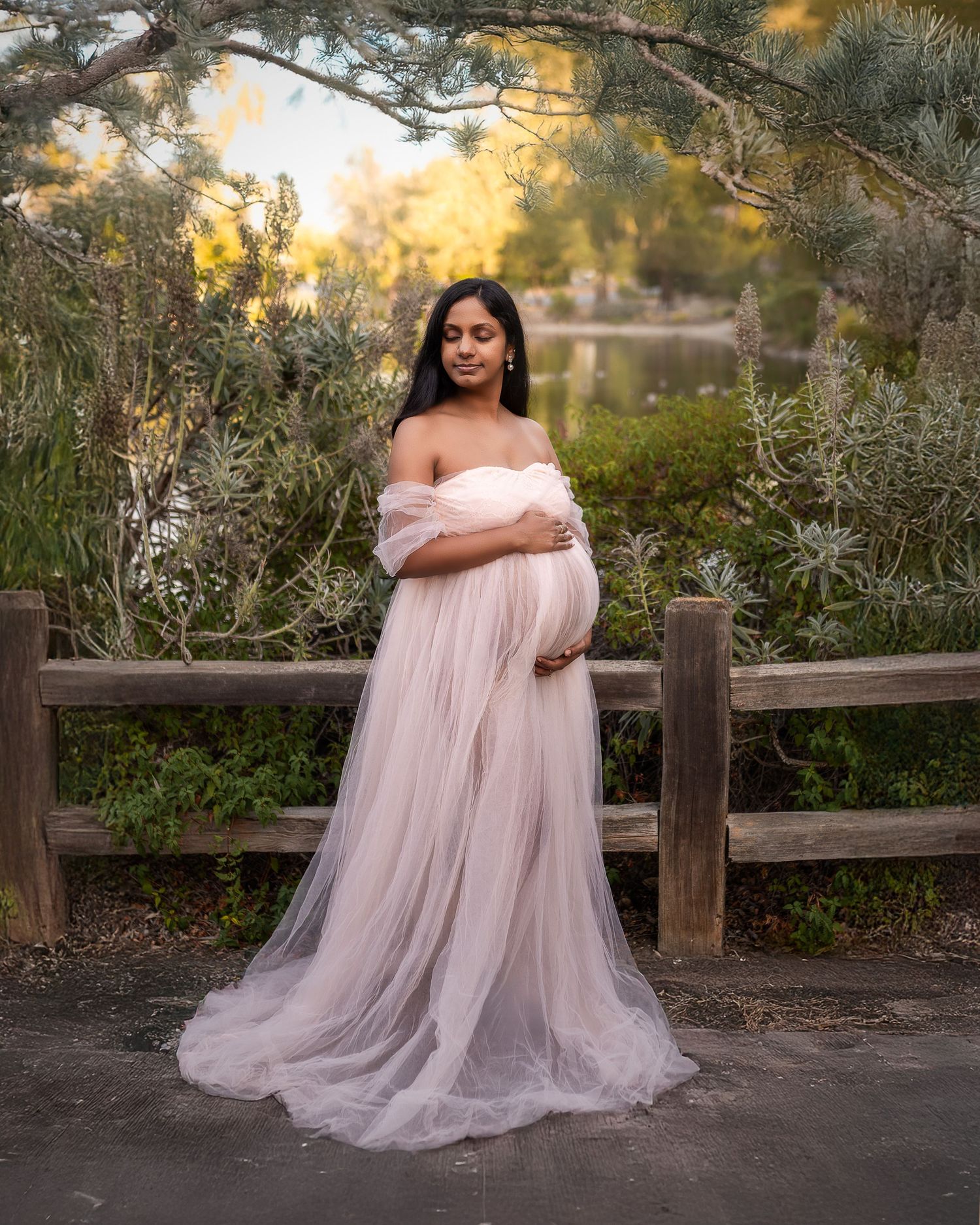 A person in a flowing pink tulle gown poses gracefully on a wooden fence at sunset in a natural outdoor setting.