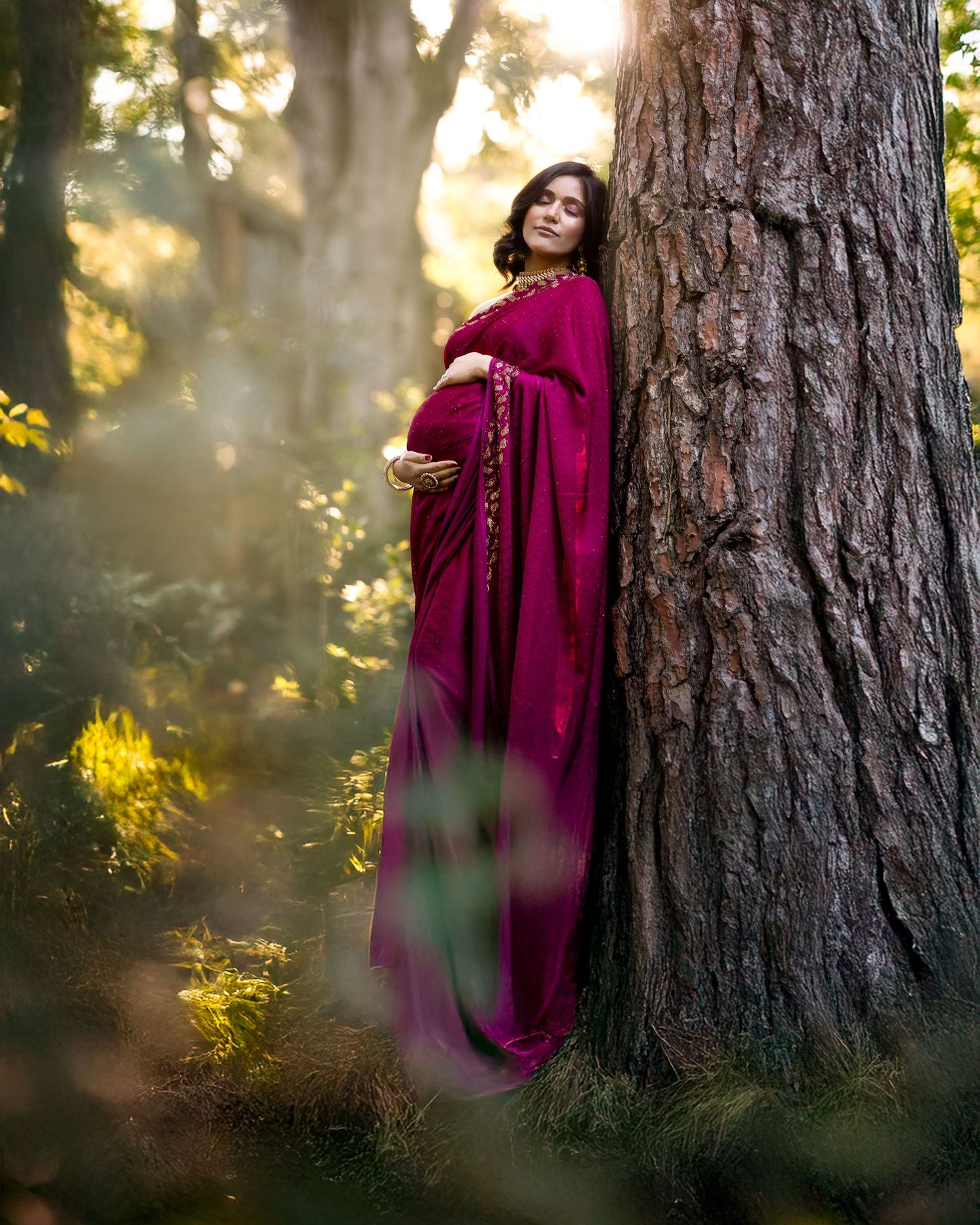Ethereal maternity photoshoot in a forest with sunlit trees and flowing burgundy dress.
